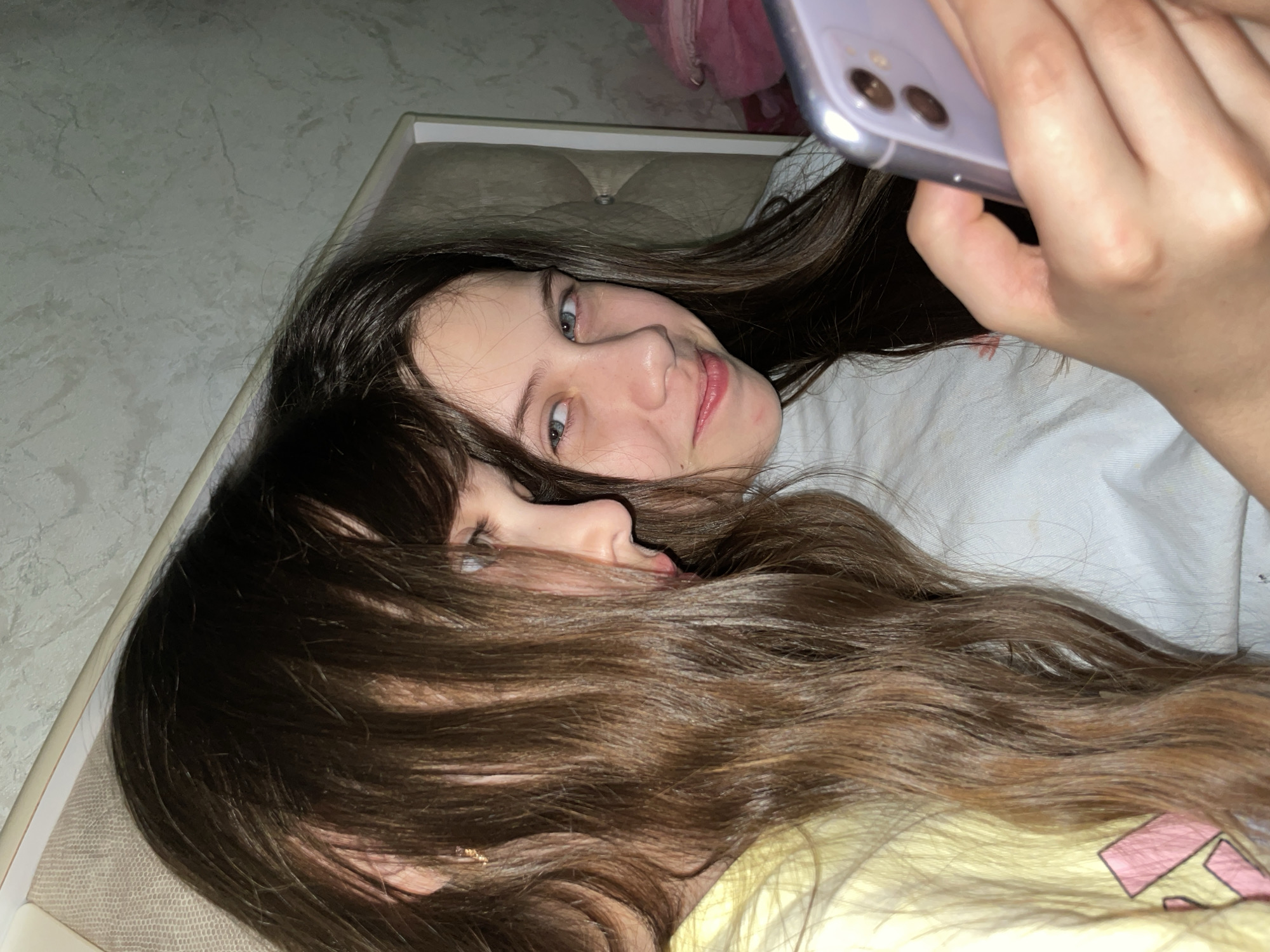 This image captures two young females indoors, likely in a bedroom or living room in Izhevsk, Russia. The shot is taken from a slightly elevated perspective, with both subjects lying down.
One female, positioned on the right side of the frame, has long, dark brown hair spread around her head. She looks directly towards the viewer with a subtle smile, her blue eyes visible. She is holding a light purple smartphone in her right hand, suggesting she is either taking a selfie or looking at the screen. She is wearing a light-colored top, possibly white or light grey.
The second female is to her left, with much of her face and body obscured by her own voluminous, long dark brown hair, which covers the lower-left portion of the image. Only a portion of her face, including an eye and her nose and mouth, are peeking through her hair. She appears to be wearing a yellow garment with a pink graphic design visible on the bottom left.
The background shows a light-colored, textured wall, possibly wallpaper, and a padded headboard or cushion of a bed or couch. The overall ambiance is casual and relaxed. No text is visible in the image.