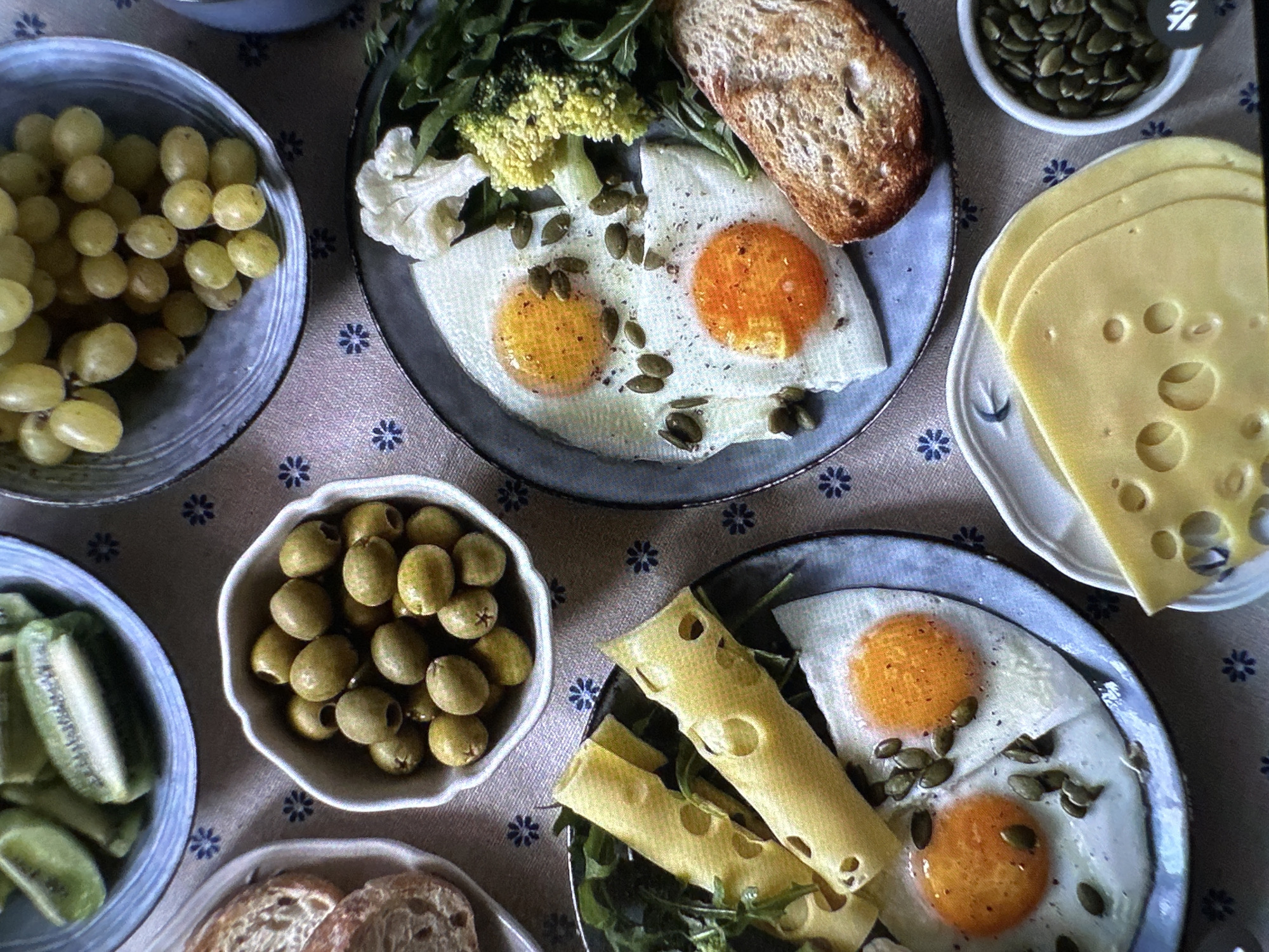 A high-angle shot shows a breakfast spread on a patterned tablecloth, not explicitly indicating Yerevan, Armenia. The arrangement includes several plates and bowls. A bowl of green grapes and a bowl of sliced kiwi are visible. Another bowl contains green olives. Two plates feature fried eggs, garnished with pumpkin seeds, accompanied by greens, cauliflower/broccoli, and slices of bread. A separate small bowl holds more pumpkin seeds. Slices of Swiss cheese are arranged on a small plate. Some cheese slices have been shaped into rolls and placed on one of the plates with the eggs.