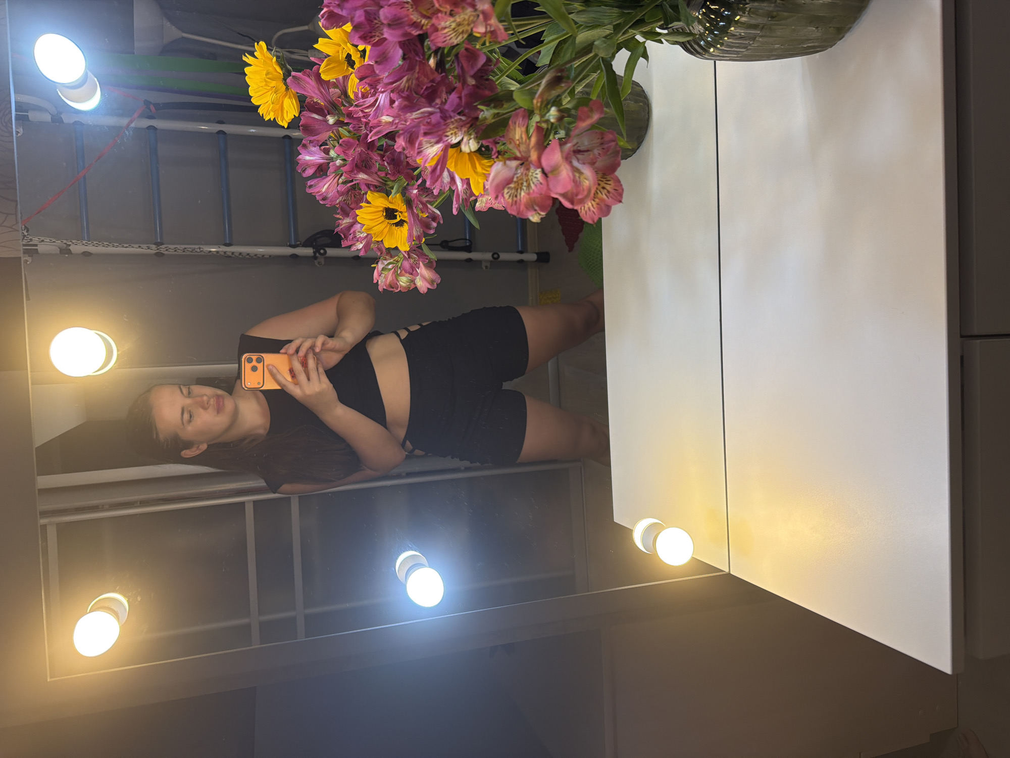 The image, rotated 90 degrees counter-clockwise, portrays a woman taking a mirror selfie in an indoor setting, likely a vanity or dressing room in Sochi, Russia. A young woman with dark hair and a calm expression is visible in the large mirror's reflection, holding an orange smartphone with multiple rear cameras, seemingly capturing her own image. She wears a black top and black shorts featuring decorative cut-outs on the sides.
The mirror frame is equipped with circular LED bulbs, some emitting a warm yellow light and others a brighter, cooler white light, illuminating the scene. To the right of the mirror, a vibrant bouquet of flowers, including bright yellow sunflowers and pink and purple alstroemeria lilies, is arranged in a textured, clear glass vase. The vase rests on a clean white surface, next to which a white cabinet or wall panel is visible.
In the background reflected within the mirror, behind the woman, an exercise area is suggested by a horizontal bar ladder with blue rungs and a white frame. Several colorful resistance bands (green, purple, and red) are hanging from this ladder, along with a red strap. Further back in the reflection, a window or similar framed structure with horizontal and vertical dividers can be seen. The time of day is not explicitly indicated but the reliance on artificial lighting suggests it might be evening or a dimly lit room. No visible text is present in the image.