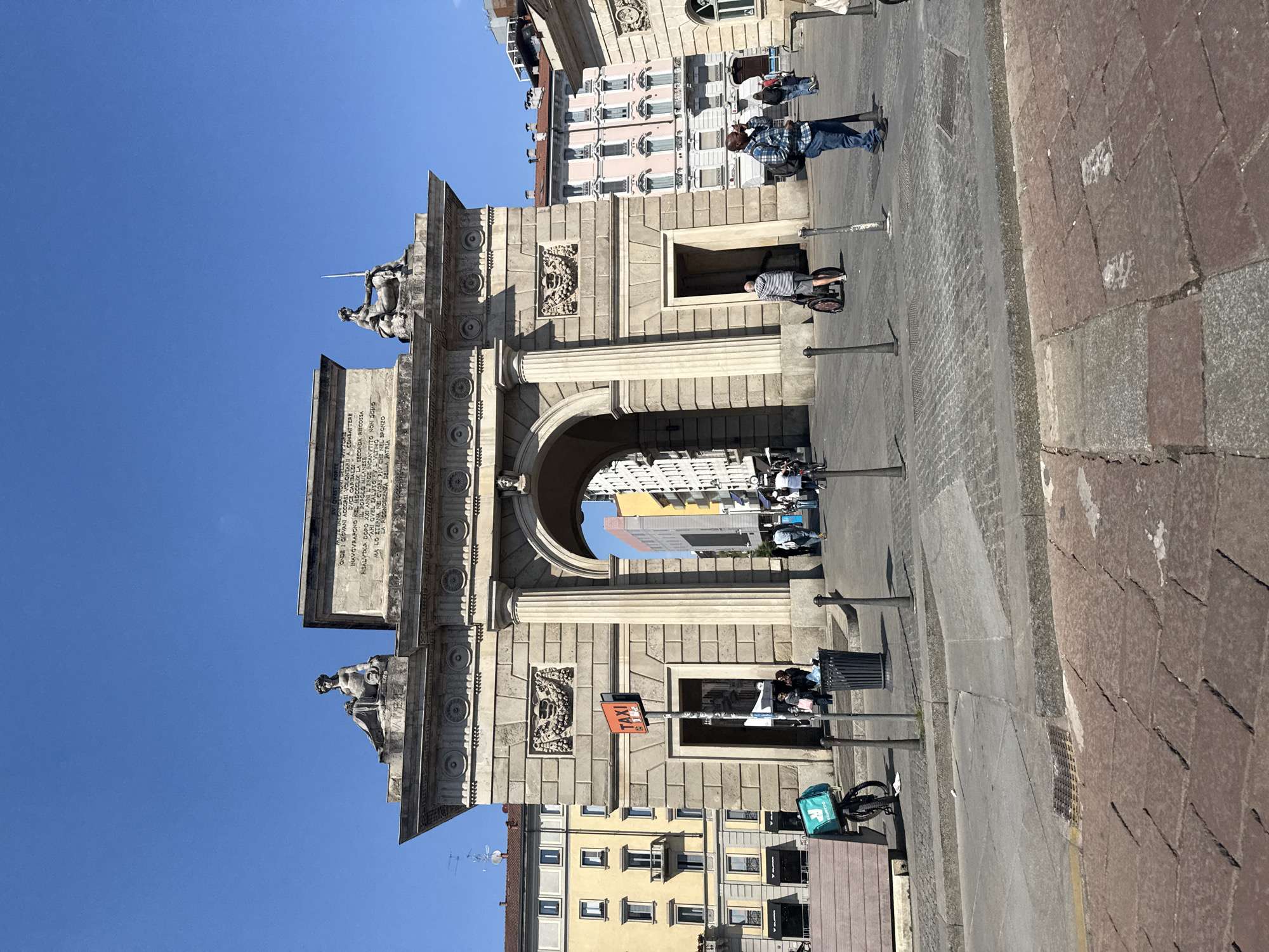 A grand stone archway, identified as part of the Porta Garibaldi in Milan, Italy, dominates the sunny urban scene. The monument, rendered in light-colored stone, features classical architectural elements, including large columns, intricate carvings, and sculpted figures atop its structure. One statue appears to be a soldier holding a spear. An inscription in Italian is visible on the upper part of the arch, with decipherable words like "GIOVANI ACCORSI VOLONTARII A COMBATTERE DUCE GARIBALDI," "POPOLO MILANESE," and "LA RICONOSCENZA DELLA PATRIA" (gratitude of the homeland), suggesting a historical commemorative purpose.
The archway provides a passage through which more city buildings can be seen. On the right side, a prominent red and orange "TAXI" sign indicates a taxi stand. Several individuals are present: a person in a plaid shirt and jeans stands near the arch, another is partially visible within an alcove of the structure, and others are further in the background. In the foreground, a bicycle with a distinctive teal delivery bag (likely Deliveroo) is parked, signaling modern urban life. The ground is paved with stone, showing a mix of cobblestones and larger slabs. The clear blue sky and strong shadows suggest a bright, sunny day.