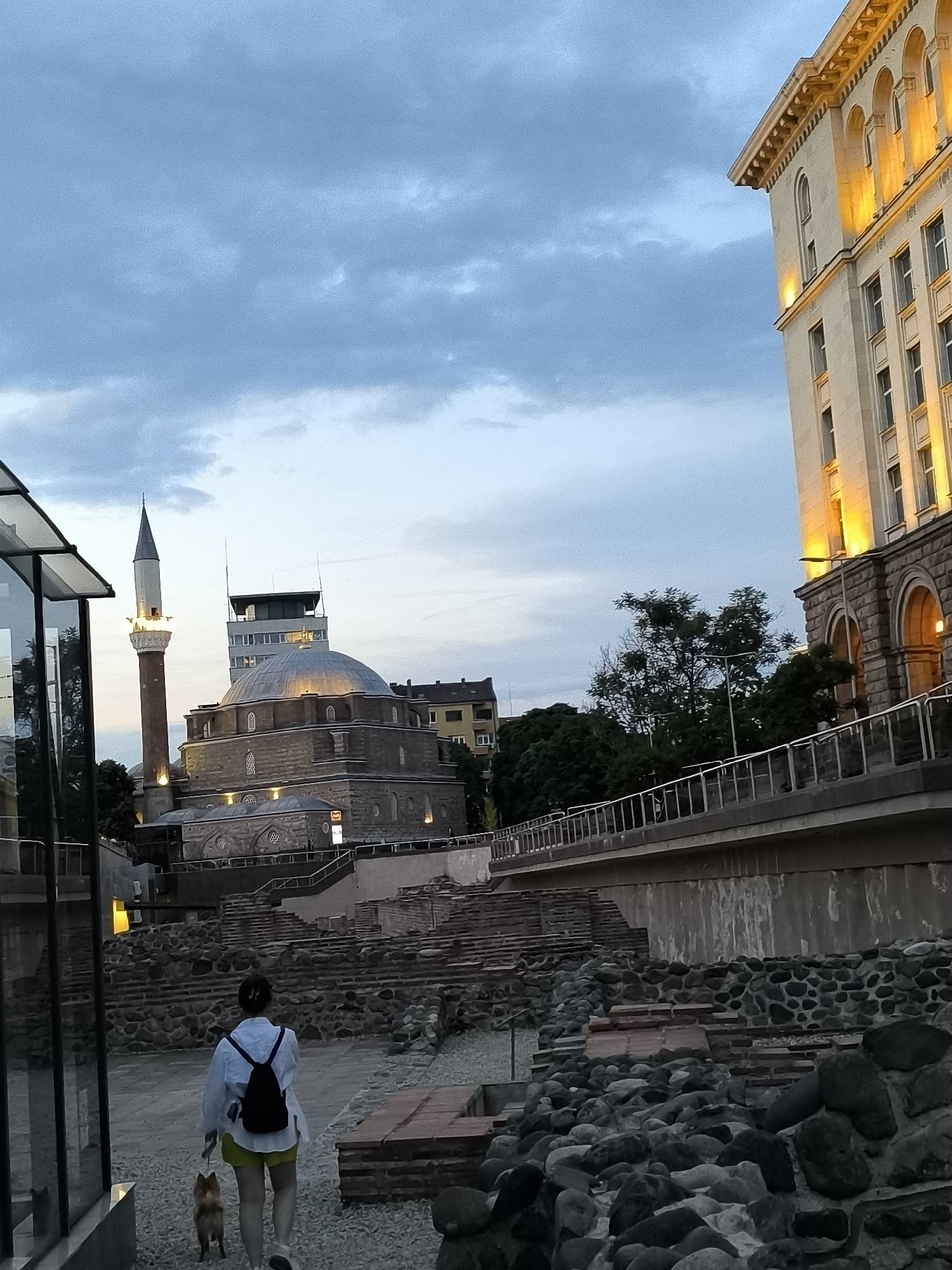The image depicts an evening scene in the city of Sofia, Bulgaria, specifically focusing on the ancient Serdica complex. In the foreground, a woman with her back to the camera is walking a small, ginger-colored dog on a leash. She is wearing a white collared shirt, yellow shorts, and a black backpack. The ground is covered with gravel and interspersed with ruins of ancient stone structures.
Behind the woman, the dominant structure is the Banya Bashi Mosque, characterized by its dome and minaret, illuminated by spotlights. Adjacent to the mosque, a tall, modern building stands, with its upper floors featuring arched windows lit with a warm, yellow light. A bridge-like structure with a metal railing runs across the midground, separating the ancient ruins from the buildings. Trees and foliage are visible throughout the scene, partially obscuring some of the background elements. The sky is overcast with a twilight ambiance.