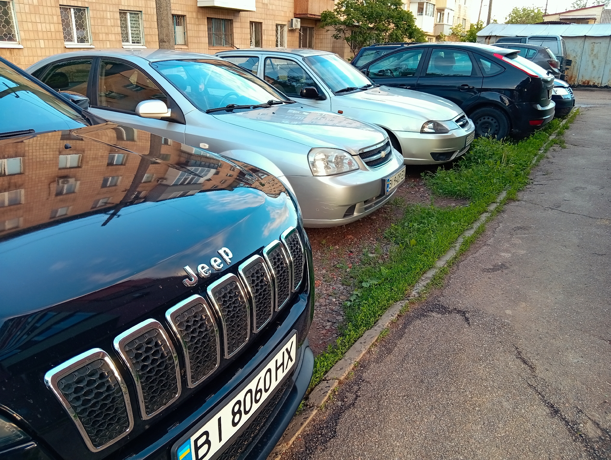 The image displays a line of cars parked on an asphalt surface alongside a grassy verge, with a residential building in the background. In the foreground, a black Jeep SUV is prominent, showing the "Jeep" emblem on its hood and a Ukrainian license plate "BI 8060 HX". Next to the Jeep is a silver sedan, identifiable by its Chevrolet grille and a Ukrainian license plate "BI 9409 EX". Further down the line, a black hatchback (possibly a Ford Focus) and other vehicles are parked.
The cars are stationary, indicating a parking scene. The reflections of the nearby building are clearly visible on the hoods and windows of the cars, suggesting a shiny surface and a bright environment. The background features a multi-story building with a light brown or tan brick facade and several windows. Green grass lines the edge of the asphalt pavement, which shows some cracks. A tree is also visible behind the parked cars.
The time of day appears to be late afternoon or early evening, characterized by the warm, directional light and the shadows cast. The weather is clear and sunny. The scene provides a glimpse into a typical street or parking area in an urban or suburban setting. The Ukrainian license plates, specifically with the "BI" prefix, confirm the location as the Poltava region of Ukraine. No individuals are visible in the image.