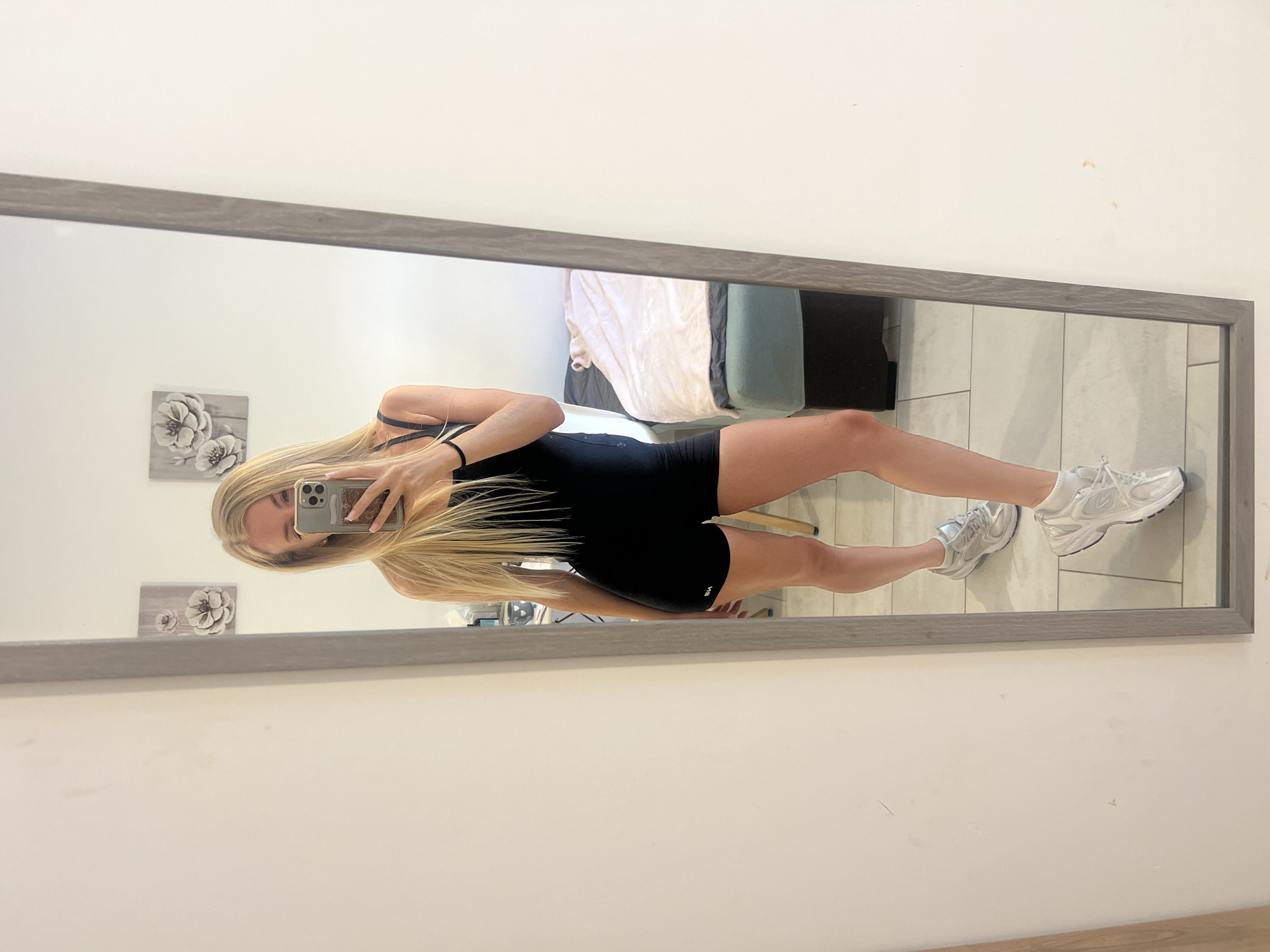 A young woman with long blonde hair is taking a mirror selfie in a room. She is wearing a black athletic bodysuit and white sneakers. The room appears to be a bedroom, with a bed and two framed floral pictures on the wall. The mirror is tall and has a gray wooden frame. The floor is tiled. There is a bed with pink and white bedding visible in the reflection of the mirror. The scene is well-lit.