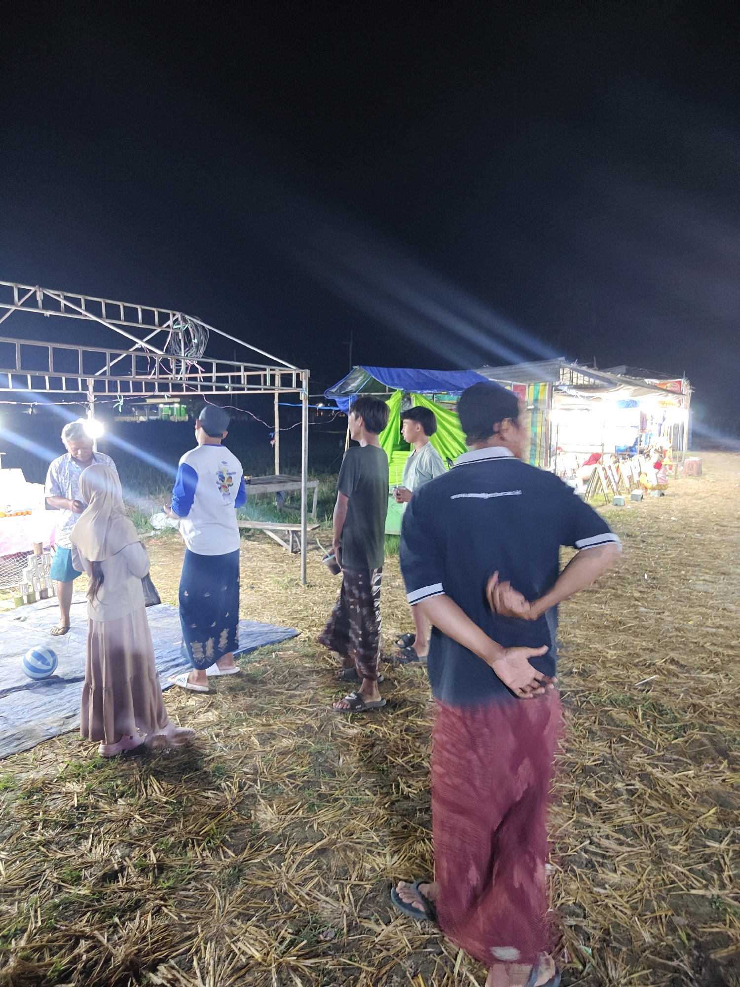 At night in Suling Wetan, Indonesia, a group of people stand in a field of dry grass. Several individuals are gathered near a temporary structure with a metal frame and fabric coverings, possibly a market stall or event tent. One person in the foreground wears a dark t-shirt and a maroon sarong, with their hands clasped behind their back. To their left, a person in a white long-sleeved shirt and a patterned sarong faces away from the camera. In the middle ground, a woman wearing a light brown hijab and dress stands on a blue tarp, near a striped ball. Other individuals are visible in the background, including two men standing by the tent, one in a green shirt and the other in a light blue shirt. The scene is illuminated by overhead lights and spotlights, casting long shadows. The environment appears to be an open outdoor area, likely a gathering or festival.
