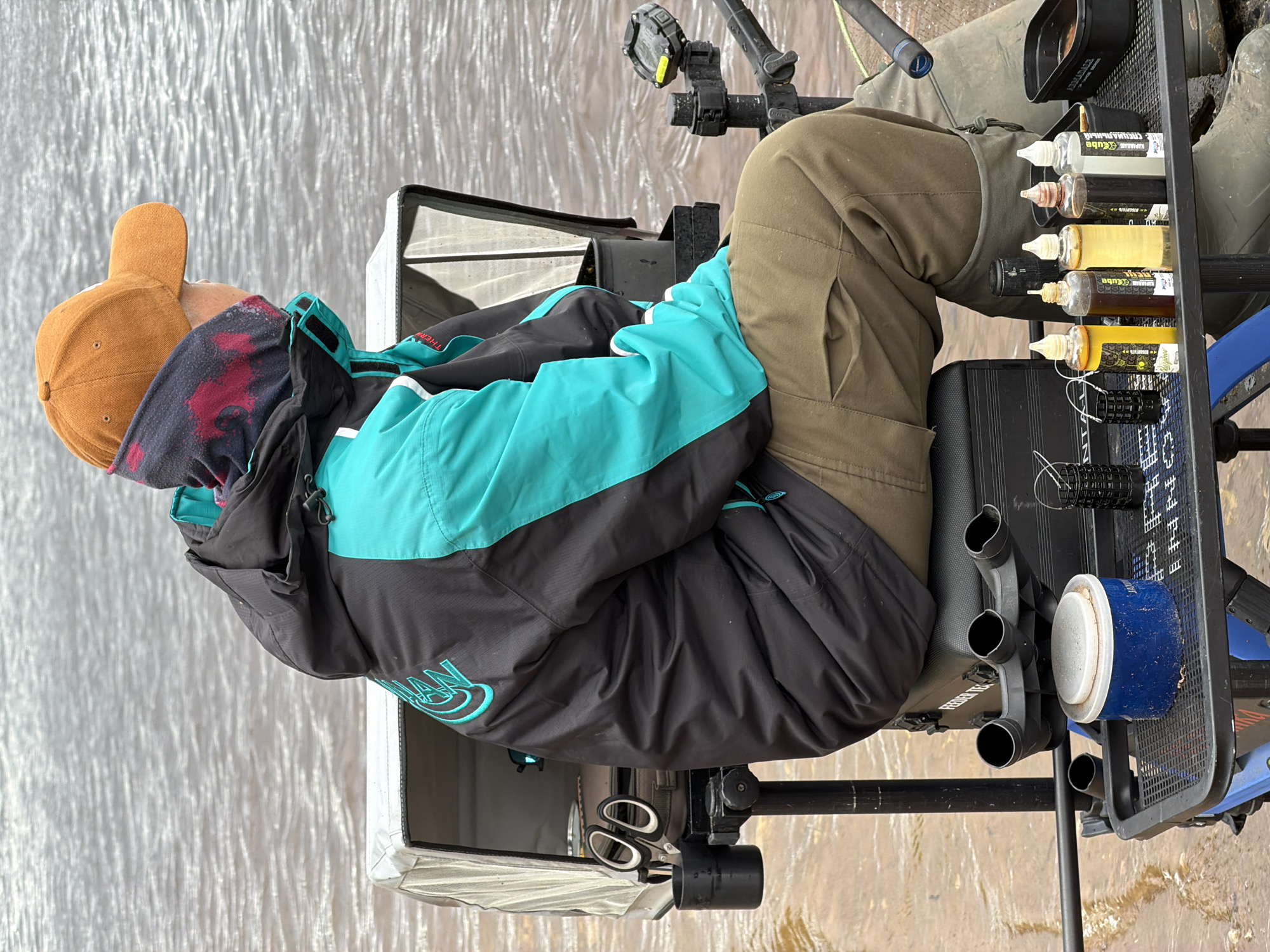 An individual is depicted from the side, facing right, seated on a black fishing platform at the edge of a body of water, which is assumed to be in Chaikovsky, Russia. The person is wearing an orange baseball cap, a multi-colored neck gaiter with red accents, a black and teal/aqua two-tone jacket with a partially visible teal logo on the back, olive green cargo pants, and dark green rubber boots.
The fishing setup includes a black cup attached to the left of the seat, containing a pair of silver scissors with white-handled grips. A white mesh-sided container is positioned behind the person. Below the seat, a black rod rest with three cylindrical openings is visible. A black perforated tray in front of the person holds a blue cylindrical container with a white lid, a small black wire feeder basket, and a row of five translucent squeeze bottles containing liquids of varying colors (yellow, dark amber, light amber, dark brown, and off-white), some with partially legible labels. To the right, a section of a fishing rod is visible, along with another rod holder supporting an electronic device on its top. The fishing platform itself has the text "FEEDER TEC" visible on its side.
The environment consists of a rippled, brownish body of water in the background, suggesting a river or large lake. The ground in the immediate foreground appears wet and muddy where the fishing station is set up.