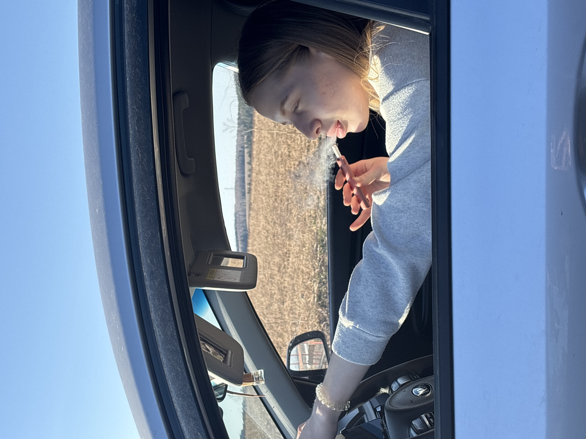A fair-skinned young woman with light brown hair leans out the window of a silver car. She is wearing a light gray sweatshirt and a gold bracelet on her right wrist. She is holding a thin, pink object in her right hand and exhaling a vapor, possibly from an electronic cigarette or vape device. The car is parked in a rural area with a field of dry, brown vegetation visible in the background. The sky is clear and blue. The interior of the car is visible, including the steering wheel and sun visor. The exact location is unknown.