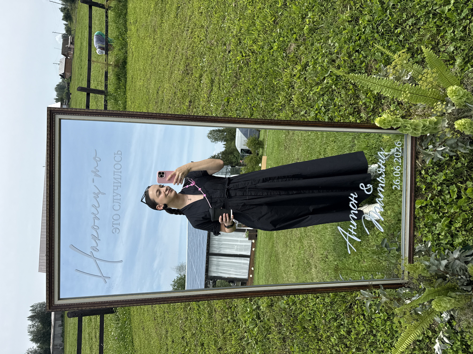A woman in a black dress is taking a selfie in a large, ornate mirror placed outdoors. The mirror reflects her and the surrounding environment. In the mirror, text is visible at the top and bottom. The top text in cursive appears to read "Наконец-то это случилось." The bottom text, also in cursive, reads "Антон & Яна" and a date "26.06.2026". The woman is holding a black cup in her left hand and a pink smartphone in her right hand. She is wearing black earrings and a beaded necklace. The background outside the mirror shows a grassy field with a wooden fence, some buildings, trees, and a cloudy sky. The location is not explicitly stated as being in the city of Svetloye, Russia, but the text on the mirror is in Russian.