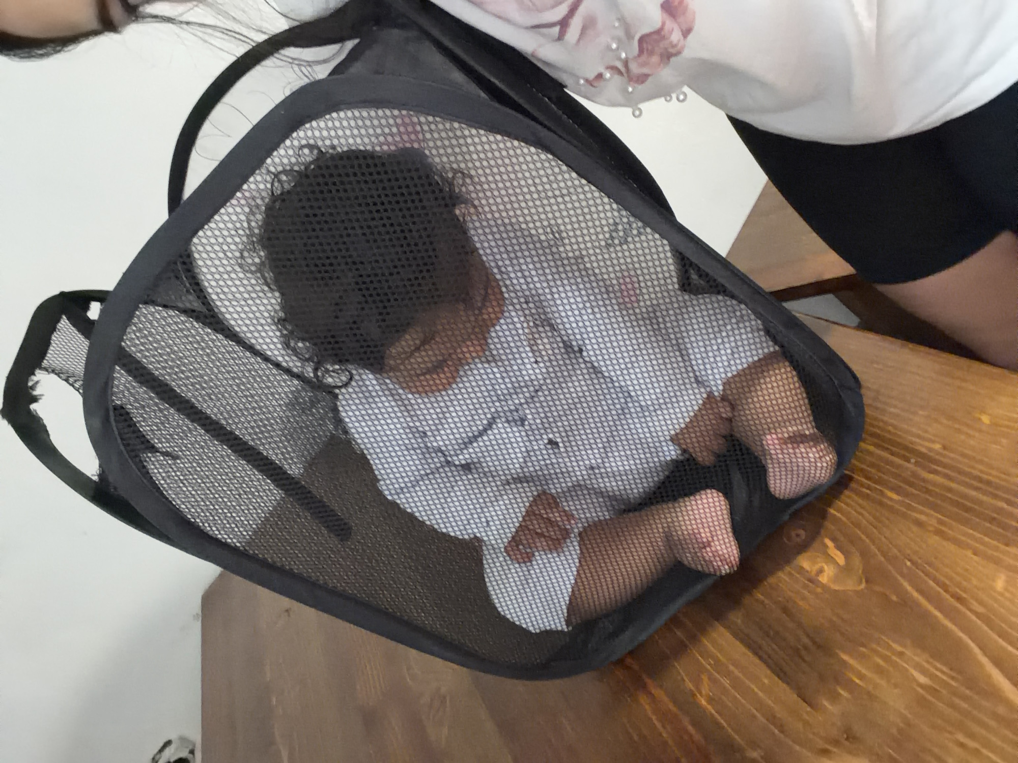 A close-up, high-angle shot shows a baby lying in a black mesh laundry basket on a wooden table. The baby is wearing a white outfit with a subtle pattern and has dark, curly hair. The baby's legs and feet are visible, resting inside the basket. A person's arm and shoulder, wearing a white shirt with a floral print and small pearl embellishments, are partially visible on the right side of the frame. The scene takes place indoors, with a light-colored wall in the background. The image is set in Razhevo Konare, Bulgaria.