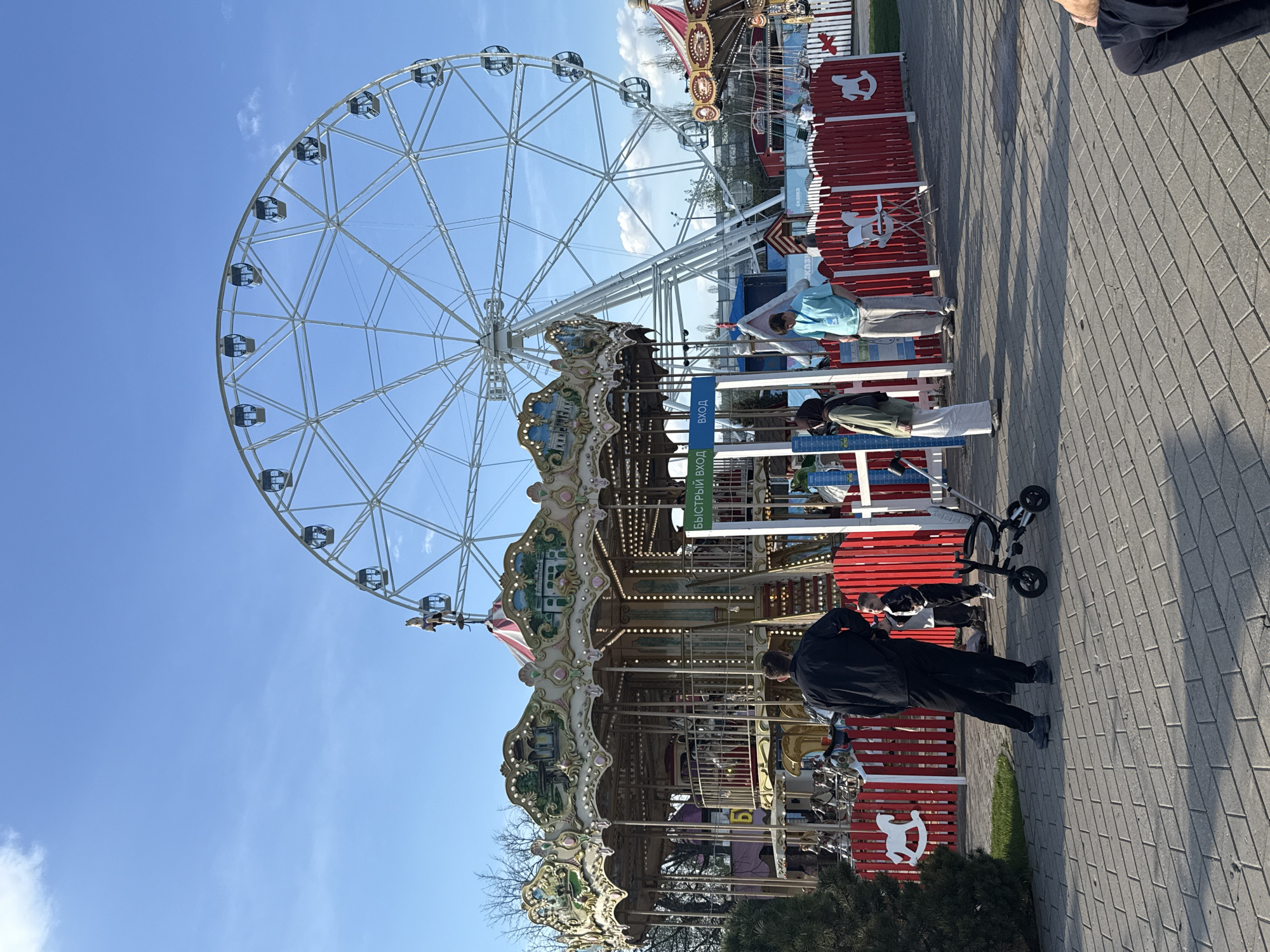 The image depicts a fairground or amusement park. A large Ferris wheel with white spokes and gondolas dominates the background. In the foreground is a carousel with ornate decorations and horses. Several people are visible, including a person in a black jacket standing near a stroller. A red fence with white horse cutouts surrounds part of the carousel area. A sign with Russian text is also visible. The location is Moscow, Russia. The sky is blue, suggesting a clear day. The ground is paved with brick or stone.