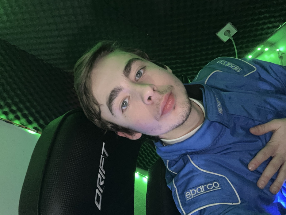 A young man with light brown hair and blue eyes poses for a selfie. He is wearing a blue Sparco racing suit and leaning against a black "Drift" gaming chair. The background features dark green soundproofing foam and green fairy lights. An electrical outlet is visible in the upper right corner. His expression is relaxed and casual.