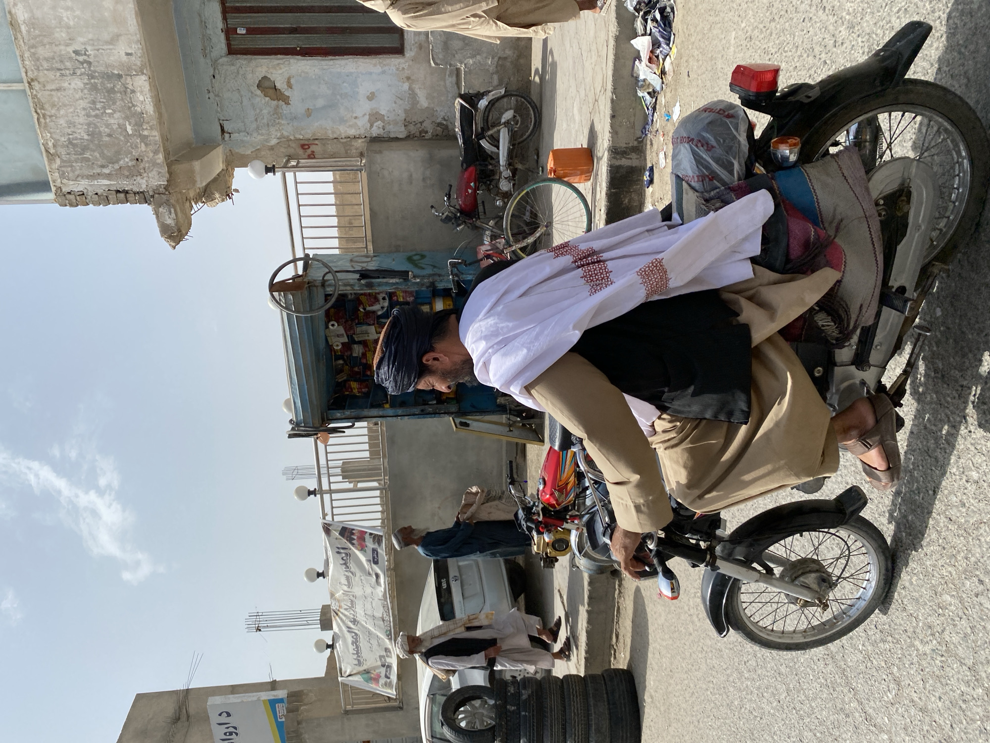 In Kandahar, Afghanistan, under a bright, hazy sky, a man wearing a dark turban and a tan tunic rides a black motorcycle. He is draped in a white cloth adorned with a red patterned border. The motorcycle is an older model with a red fuel tank and silver spokes. The man's right hand is on the handlebars, and his feet, clad in brown sandals, rest on the footpegs.
Behind the motorcycle rider, a street vendor's stall is visible, displaying various goods on shelves. To the left, two older men are walking. One, wearing a white turban and a tan vest over a light-colored robe, walks beside a white sedan. The other man, in a dark blue robe and a white turban, carries a sack.
In the foreground, a stack of black tires is piled on the side of the road. The street is paved with asphalt, and debris, including plastic bags and torn fabric, litters the ground near the curb. The buildings in the background appear weathered, with exposed brick and peeling paint, suggesting a mix of construction and disrepair common in urban environments. The overall scene depicts a typical day on a street in Kandahar, characterized by local commerce and daily life.