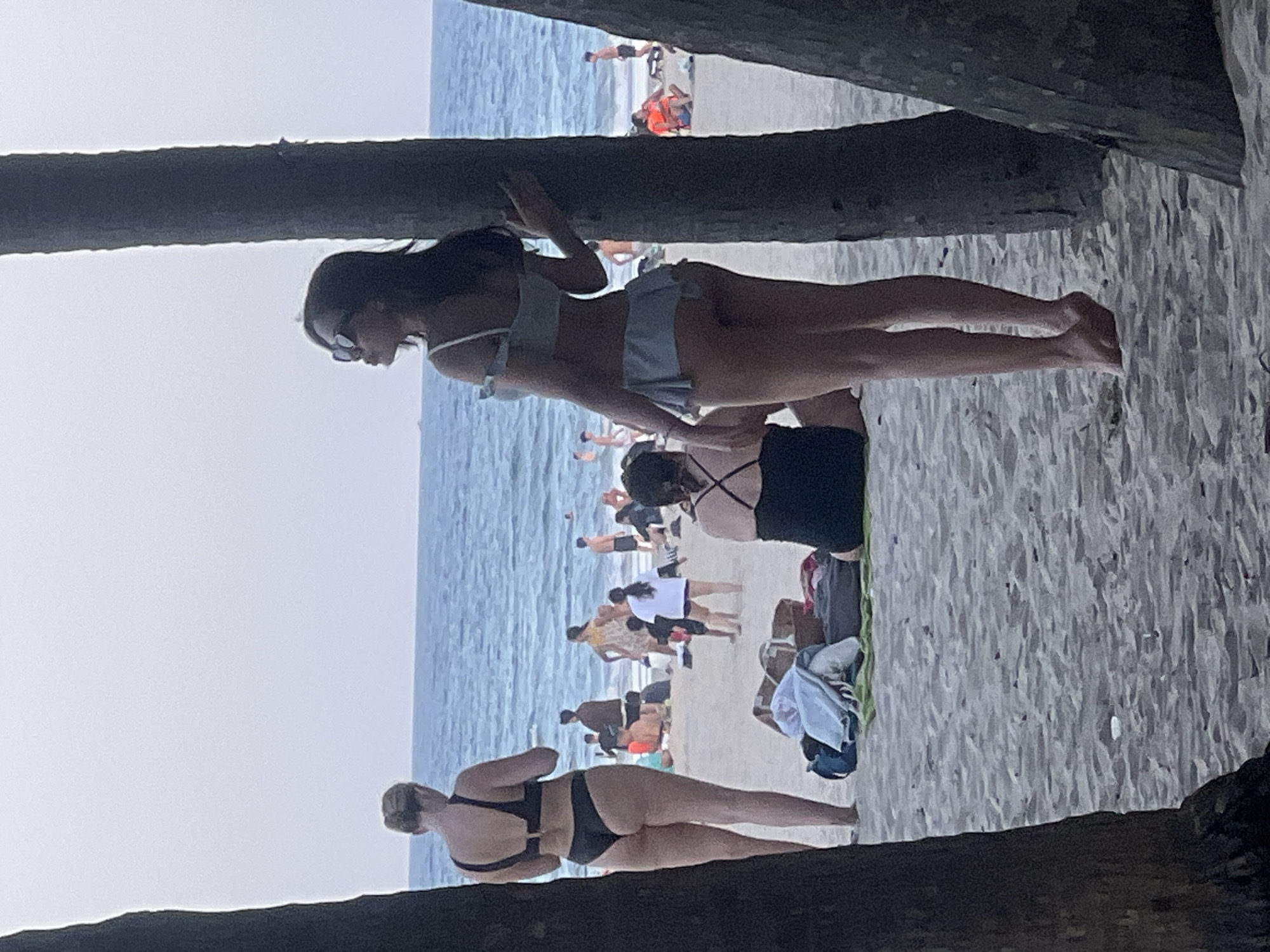 The image captures a beach scene in Da Nang, Vietnam, with the ocean and sky forming the background. In the foreground, two palm trees frame the view. On the sandy beach, several individuals are present. To the left, a woman in a black bikini stands facing away from the camera. To the right, a woman in a light-colored bikini stands with her back to the camera, facing the ocean, her hand resting on a palm tree. In the mid-ground, other beachgoers are visible, some sitting or standing on the sand. Beach towels and bags are scattered on the sand. The lighting suggests it is late afternoon or early evening.