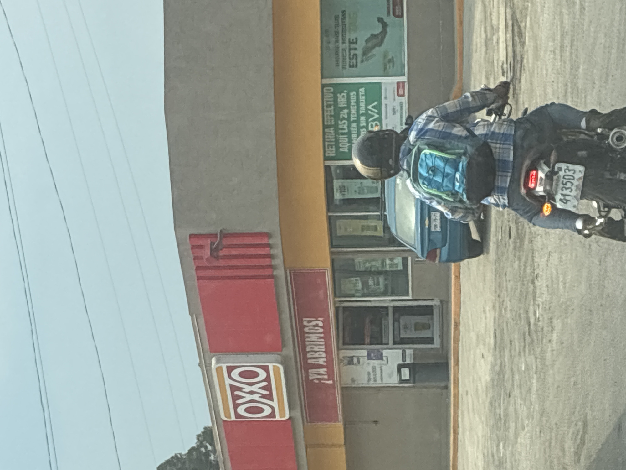 The image shows an OXXO convenience store in Cuautitlán Izcalli, Mexico. The store is yellow and gray with red signage, including the OXXO logo and the phrase "¡YA ABRIMOS!" (We're open!). There are posters in the windows, including one for BVA bank. A person on a motorcycle is visible in the foreground, wearing a helmet, a plaid shirt, and a backpack. The license plate of the motorcycle reads "413503." Overhead electrical wires are in the background. The day appears clear.