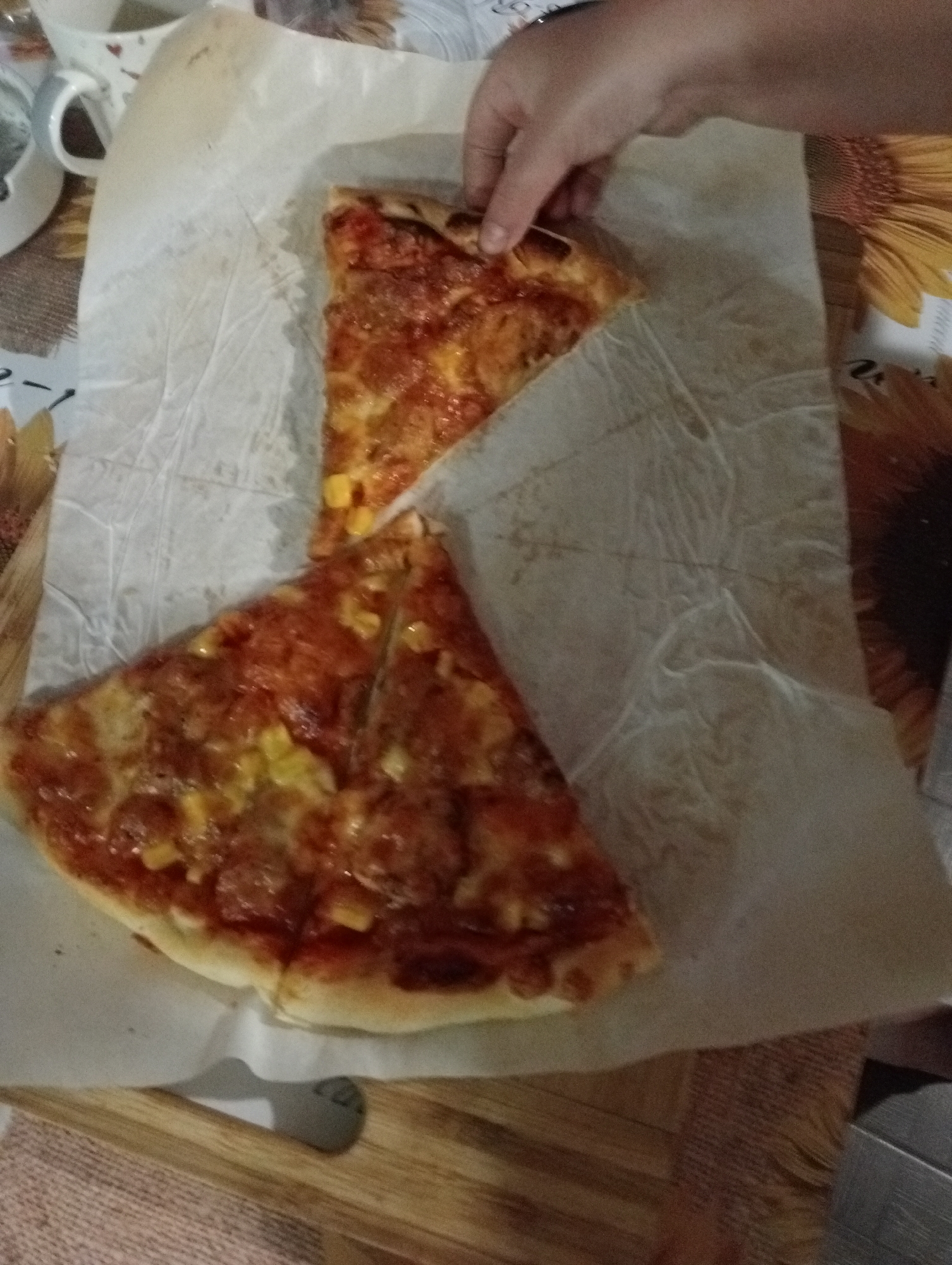 Two slices of pizza are on a piece of parchment paper. A person's hand is reaching for the top slice of pizza. The pizza is topped with sauce, cheese, and what appears to be corn. The parchment paper is on a wooden surface. In the background, there are parts of a mug and a table runner with a floral pattern.