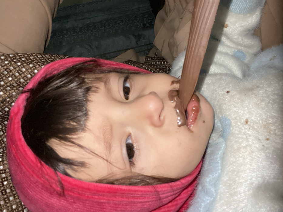 The scene is an indoor setting in Kandahar, Afghanistan. A young child is depicted in a close-up, chest-up shot. The child has dark hair with bangs visible below a magenta-colored head covering that frames their face. They are wearing a light-colored, possibly white or light blue, fuzzy or knitted sweater. The child's mouth is open, with significant amounts of melted brown chocolate smeared on the lips, chin, and surrounding facial area. A flat, elongated, dark brown object, consistent with a chocolate bar, is positioned horizontally at the child's mouth, with the child's tongue extended to engage with it. Droplets or smears of chocolate residue are visible on the child's sweater. The background consists of a brown, patterned fabric, likely upholstery, in the upper right, and a darker, possibly green or blue, draped fabric to the right, suggesting an interior environment.