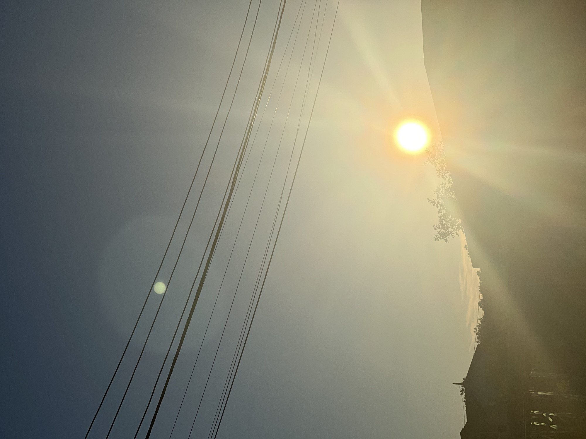 The image depicts a sunset over a suburban area. The sun is a bright orb in the lower right quadrant of the frame, casting long rays that create a hazy effect across the scene. Several horizontal power lines stretch across the upper portion of the image, bisecting the clear blue sky. Silhouetted against the bright sky are the rooftops of buildings and the tops of trees. A faint, pale circle, possibly the moon or a lens flare, is visible in the upper left quadrant. The overall impression is one of a calm, late afternoon or early evening.