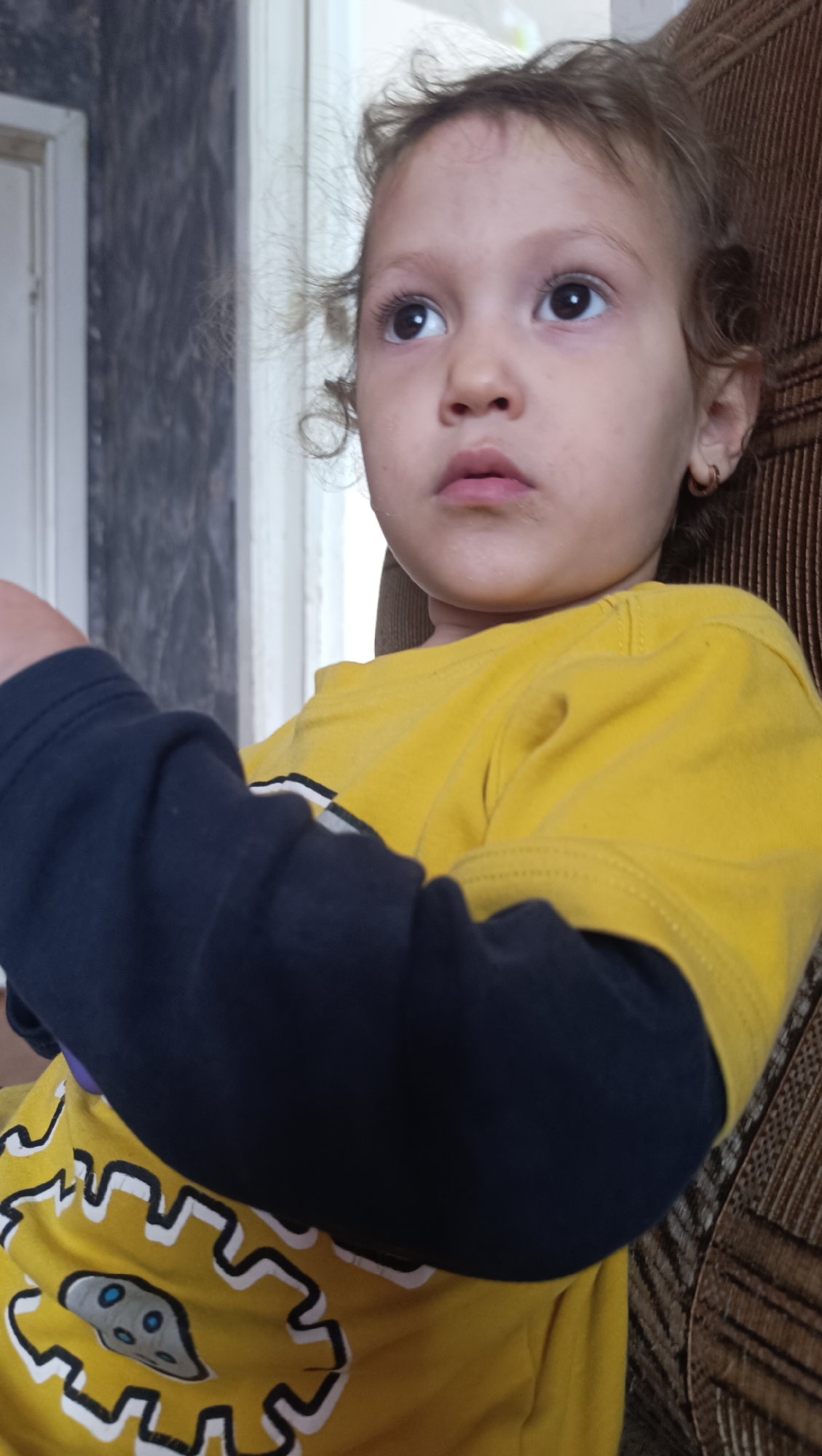 A child, approximately 3-4 years old, with curly brown hair and dark eyes, is seated on a brown upholstered chair. The child is wearing a yellow long-sleeved shirt with a dark blue long-sleeved undershirt. The yellow shirt has a graphic print featuring a white gear-like design with a grey alien-like figure with blue eyes in the center. The child's arms are crossed in front of their chest, with the dark blue sleeves visible. The background includes a portion of a door frame and a textured wall. The lighting appears to be natural. There is no indication of Karsha, Russia, or any other specific location.