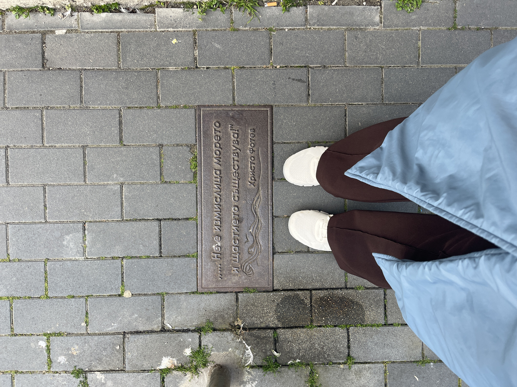 An overhead shot captures a person's lower body standing on a paved outdoor surface in Burgas, Bulgaria. The person is wearing dark brown trousers, white sneakers, and a light blue puffy jacket or coat. Their feet are positioned on the grey rectangular pavers, which are arranged in an interlocking pattern. Small patches of green grass and weeds grow in the cracks between the pavers, indicating some weathering or natural growth.
Embedded in the pavement directly in front of the person's feet is a rectangular metal plaque with Cyrillic text. The plaque is dark-colored, likely bronze or a similar alloy, and features a decorative wavy pattern below the text, possibly symbolizing the sea. The visible text reads:
"...НЕ Е ИЗМИСЛИЦА МОРЕТО
И ЩАСТИЕТО СЪЩЕСТВУВА!"
ХРИСТО ФОТЕВ
This translates to:
"...THE SEA IS NOT A FABRICATION
AND HAPPINESS EXISTS!"
HRISTO FOTEV
This is a quote from the notable Bulgarian poet Hristo Fotev. The scene is brightly lit by daylight, suggesting it is daytime, with soft light as no harsh shadows are visible. The overall impression is an urban or coastal setting, given the quote's reference to the sea and the known location of Burgas.