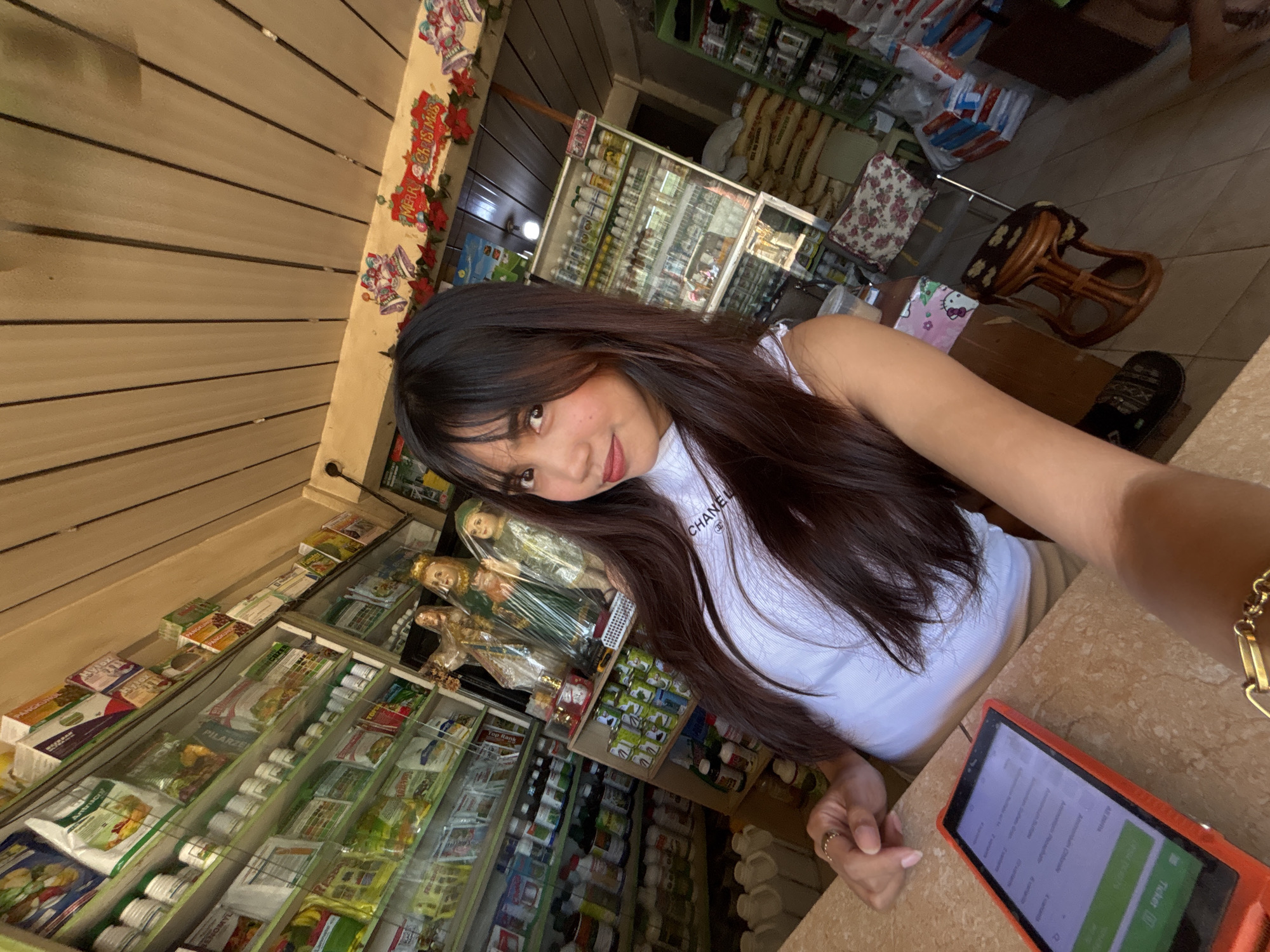 The image captures a young woman taking a selfie in what appears to be a small store or shop. The background reveals shelves stocked with various products, including bottles, packaged goods, and possibly agricultural supplies. Religious statues, specifically of saints, are visible on a shelf behind the woman, suggesting a Filipino cultural context, possibly a sari-sari store or a small general merchandise shop. Decorative "Merry Christmas" banners hang from the ceiling, indicating a festive season. The woman is wearing a white sleeveless top and is positioned in front of a counter with a tablet device. The overall lighting suggests an indoor setting, likely during the daytime. The location is confirmed to be Tagum City, Philippines, given the context of the image and its setting.