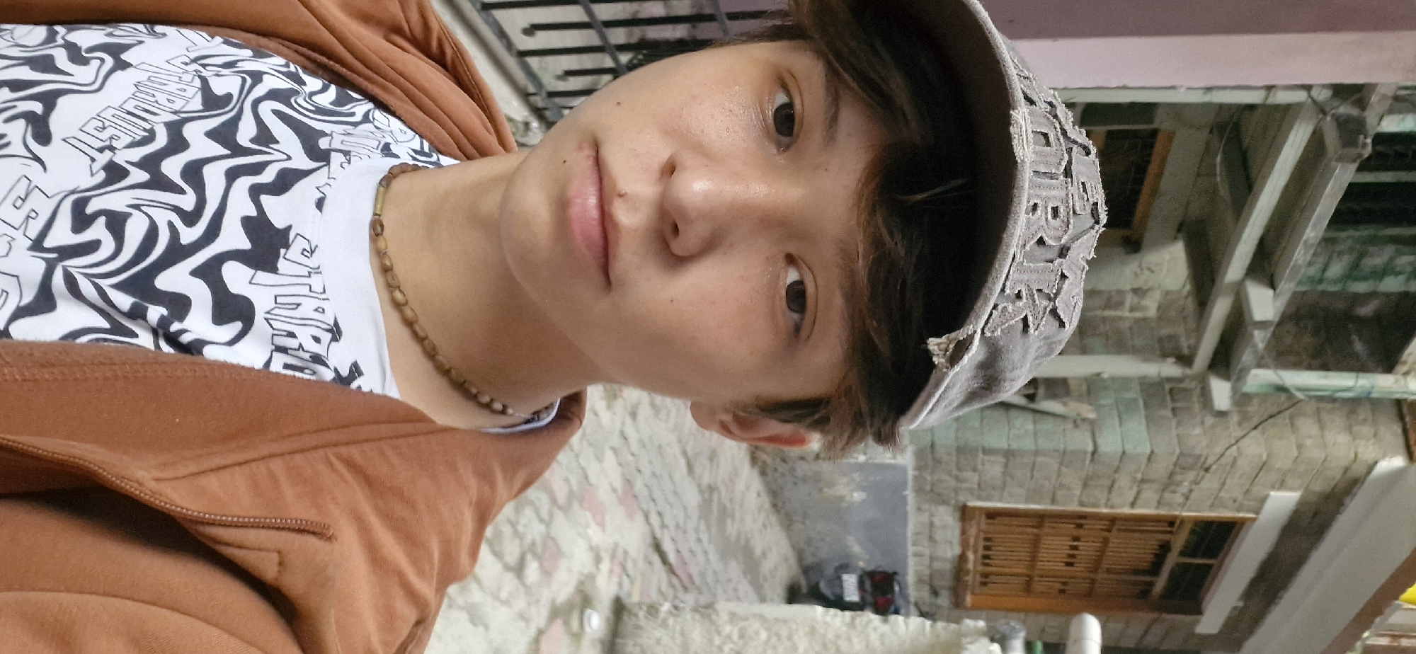 A close-up, waist-up portrait shows a young person wearing a brown cap with frayed edges and the word "YORK" in a distressed font. They have dark hair peeking out from under the cap, dark eyes, and a light complexion with a few freckles on their cheeks. They are wearing a white t-shirt with a black and white wavy pattern and text that appears to be "STARDUST" repeated in an artistic font. A brown beaded necklace with a single cylindrical bead in the center is visible around their neck. They are also wearing an unzipped, light brown hooded jacket.
The background suggests an outdoor urban environment. To the left, there is a pink-colored building with a metal railing and a roof structure supported by white beams. To the right, a stone building with wooden window frames and a visible motorcycle parked in the alleyway. The ground is paved with light-colored rectangular stones. The scene is likely in the city of Vashisht, India, given the architectural style and the stone paving. The lighting indicates it is daytime.
