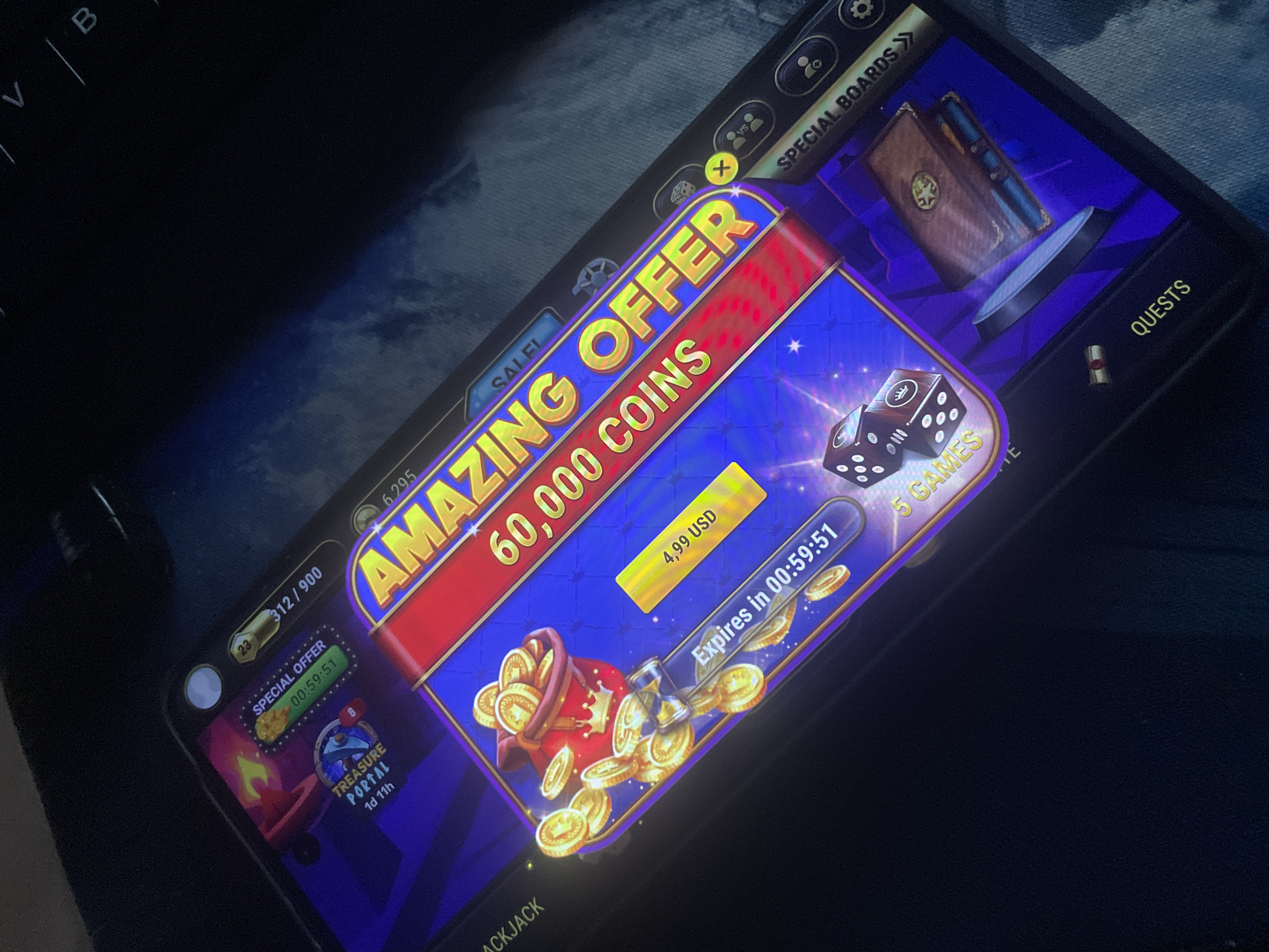 The image shows a close-up, angled view of a mobile phone screen displaying a game interface. The game appears to be a casino or gambling-themed application with vibrant graphics. Dominating the screen is a large, red and yellow banner advertising an "AMAZING OFFER" of "60,000 COINS" for "4.99 USD". Below this banner, a bag overflowing with gold coins is depicted. To the left of the offer, a timer indicates "SPECIAL OFFER" that "Expires in 00:59:51". Further to the left, a section labeled "TREASURE PORTAL" shows "1d 11h" remaining. In the lower portion of the screen, dice and text such as "5 GAMES" and "QUESTS" are visible, suggesting gameplay elements. The background is a dark, purple and blue abstract design with glowing elements. The phone is resting on a dark surface, likely a keyboard or fabric, and the overall lighting suggests an indoor, possibly dim environment. The image quality is moderate, with some blurriness and glare. There are no people or discernible location information within the image itself.
