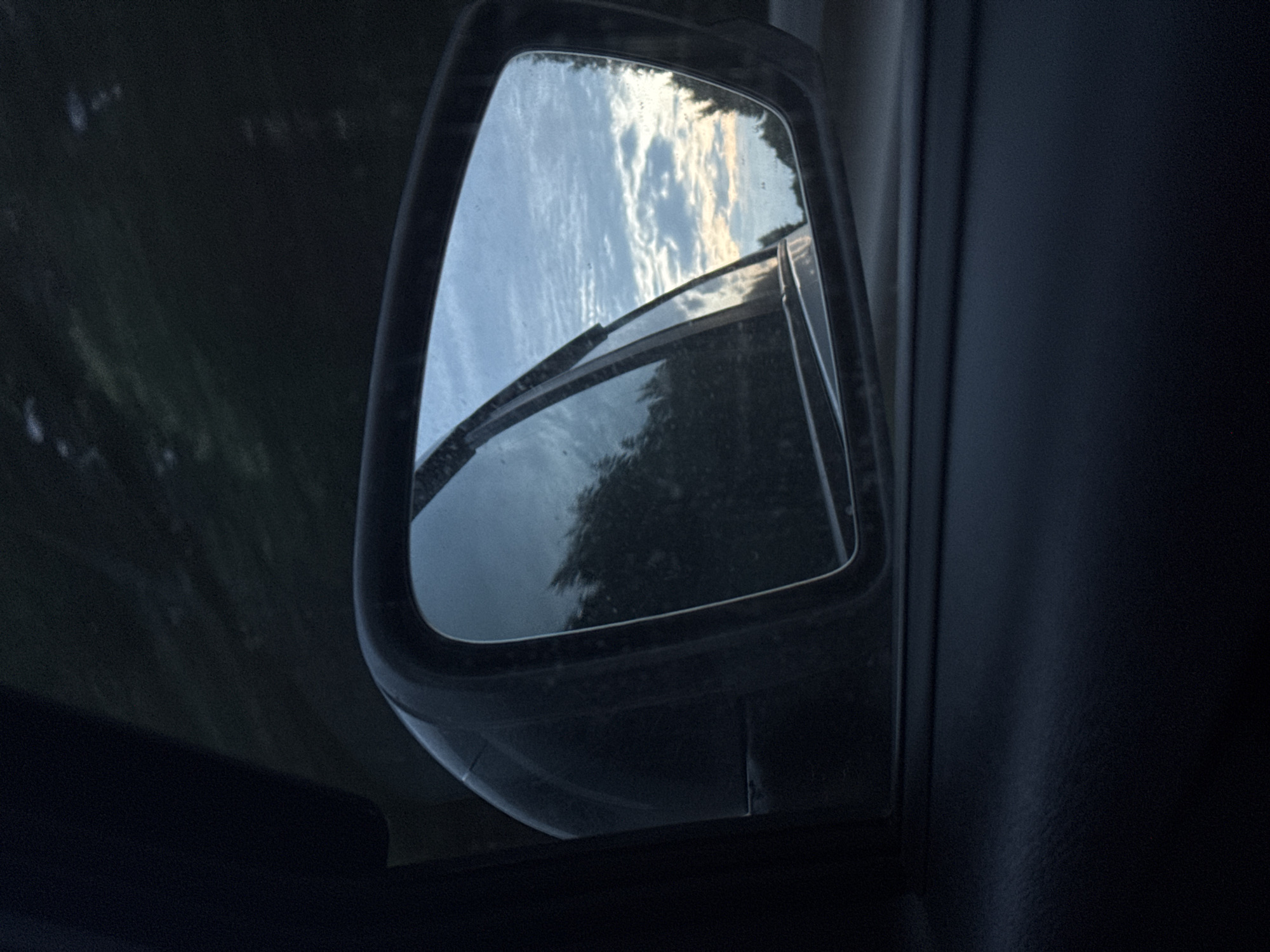 The image is a close-up shot of a car's side mirror, reflecting a cloudy sky and trees. The mirror is mounted on the exterior of the car, with the car's interior visible in the foreground to the left and bottom. The reflection in the mirror shows a dusk sky with patches of blue and white clouds, and dark silhouettes of trees. The overall lighting suggests it is either dawn or dusk. There are no people or visible animals in the image. The scene appears to be in a rural or suburban area, given the presence of trees and the overall subdued lighting. The car's mirror is slightly dirty. There is no specific information that indicates the location within Chaikovsky, Russia.