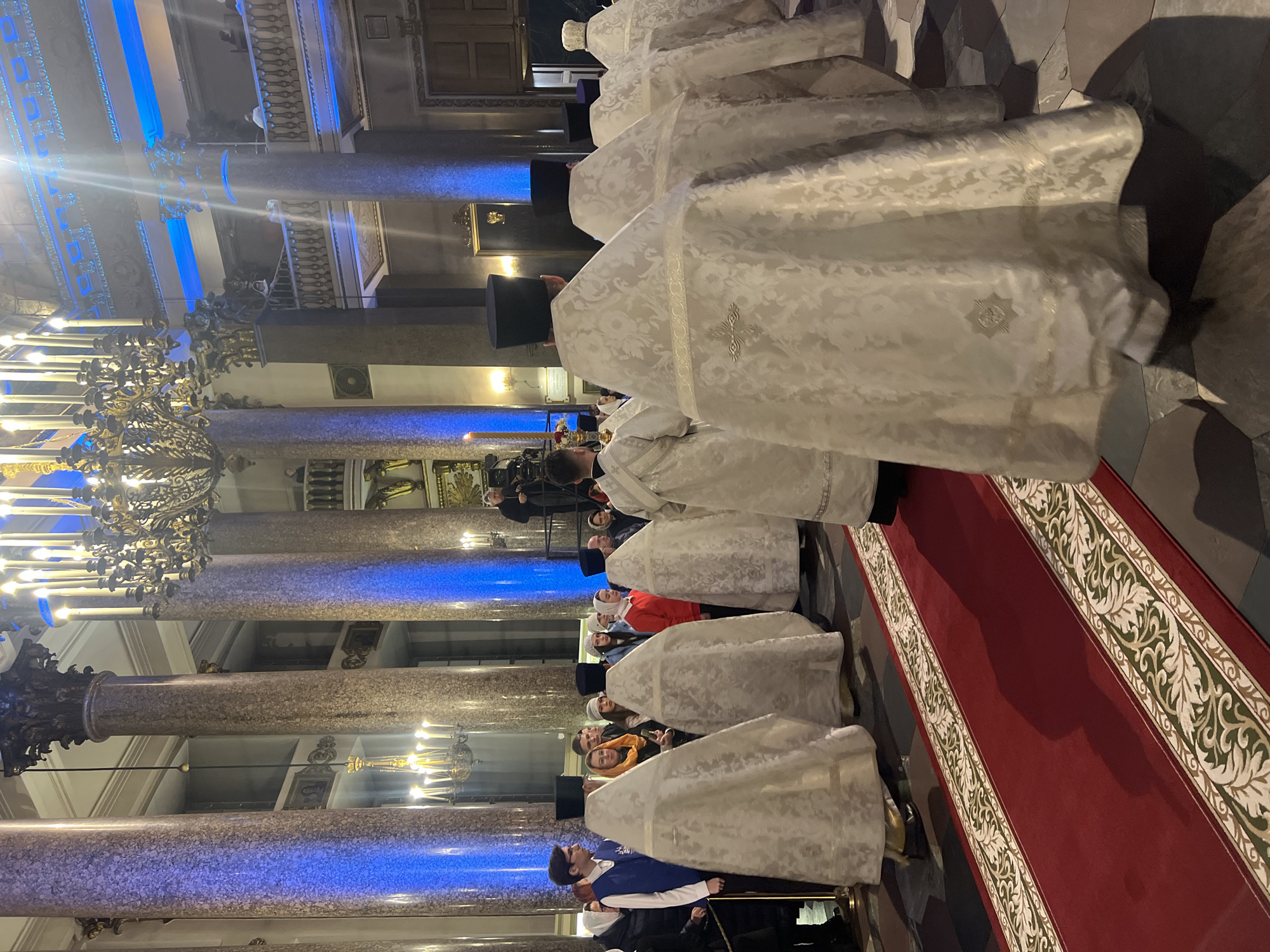 The image depicts an interior view of a church, possibly the Saint Isaac's Cathedral in Saint Petersburg, Russia, given the architectural style and general appearance of the building. The focus is on a row of people wearing white vestments with embroidered crosses on the back, suggesting they are clergy participating in a religious ceremony. The clergy are standing on a red carpet that is bordered with a white, green, and cream leaf pattern.
In the background, there are marble columns illuminated with blue light. Ornate chandeliers are also visible. Further back, additional people can be seen standing or sitting. The image is oriented vertically.