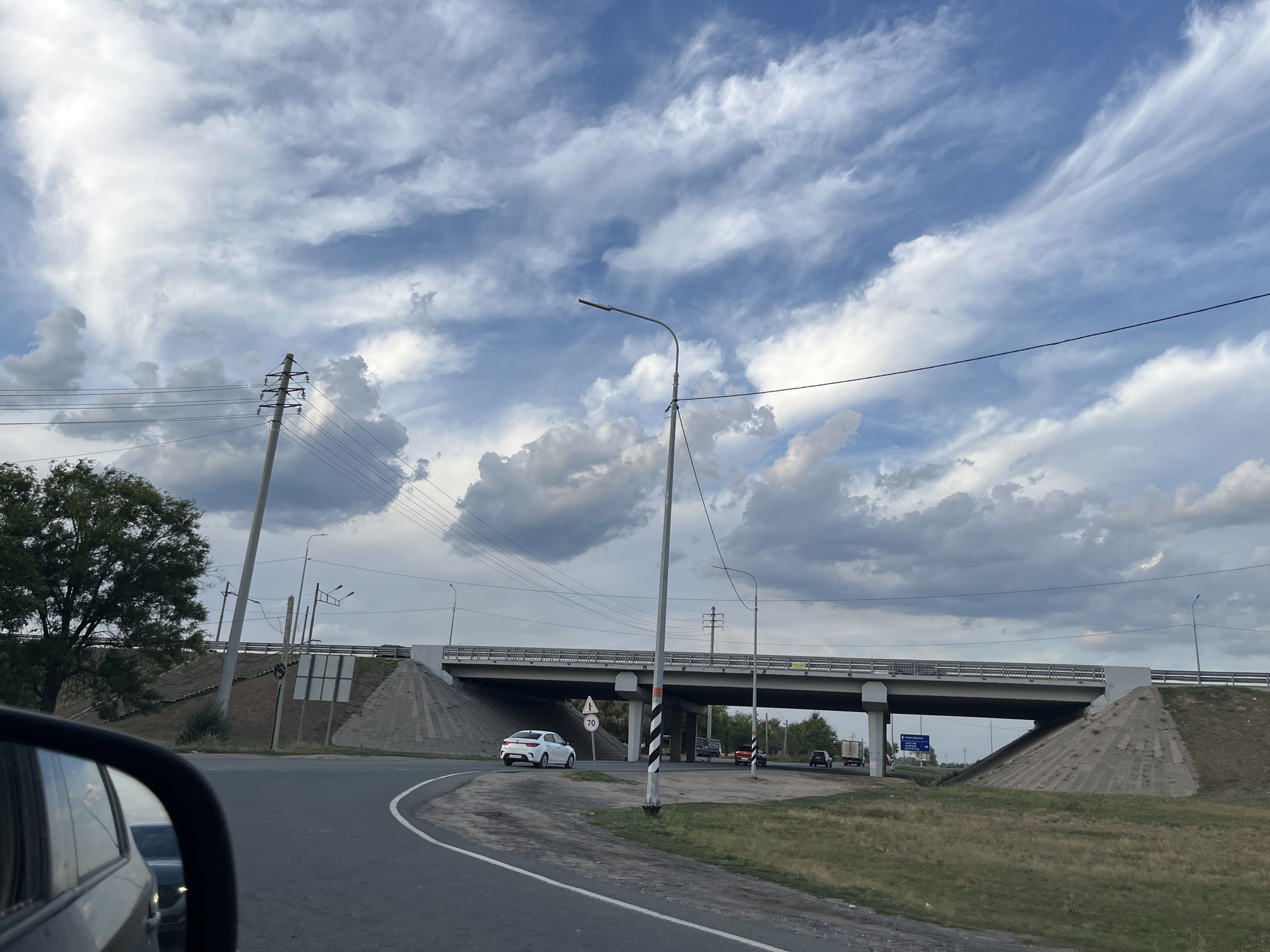 An overcast sky with dramatic, wispy clouds looms over a highway overpass in Shumeika, Russia. The overpass, constructed of concrete and steel, spans a roadway. A white sedan is visible driving under the overpass, accompanied by a speed limit sign indicating 70. Several utility poles line the sides of the road, supporting numerous wires. In the foreground, the reflection of a dark vehicle is visible in a car's side-view mirror. Green grass and trees are present on either side of the road, suggesting a semi-rural or suburban setting.