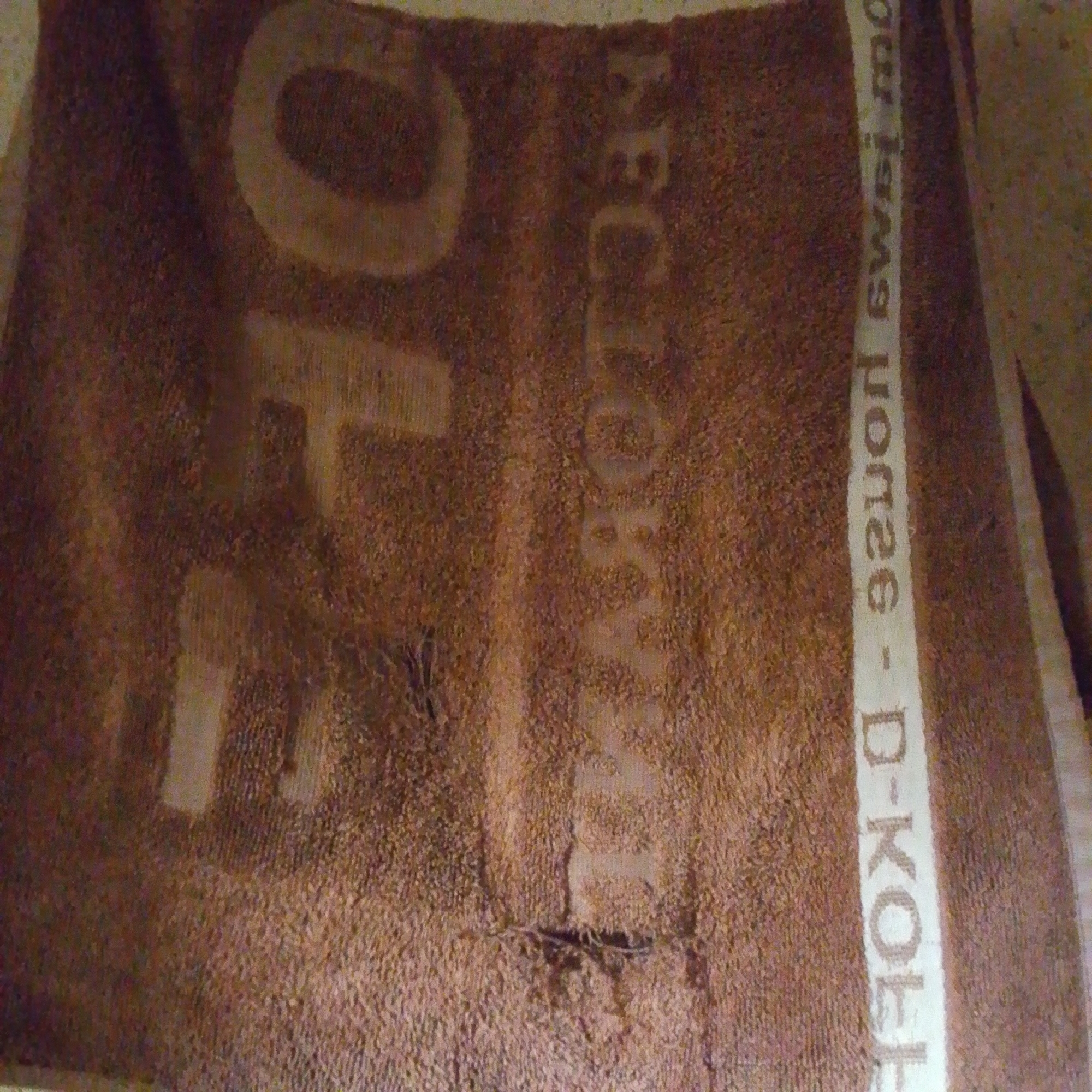 The image shows a brown towel with some text embossed on it. On the left side, the letters "FO" are visible in large print. To the right of this, running vertically, is a white stripe with text on it. The text on the white stripe appears to be in multiple languages and reads "ou isms norge - D-KOBB" and further down "eclo". There are also some holes or worn areas in the towel. The lighting suggests an indoor setting with a possibly textured floor visible in the background. No specific location in Jalingo, Nigeria, can be determined from the image.
