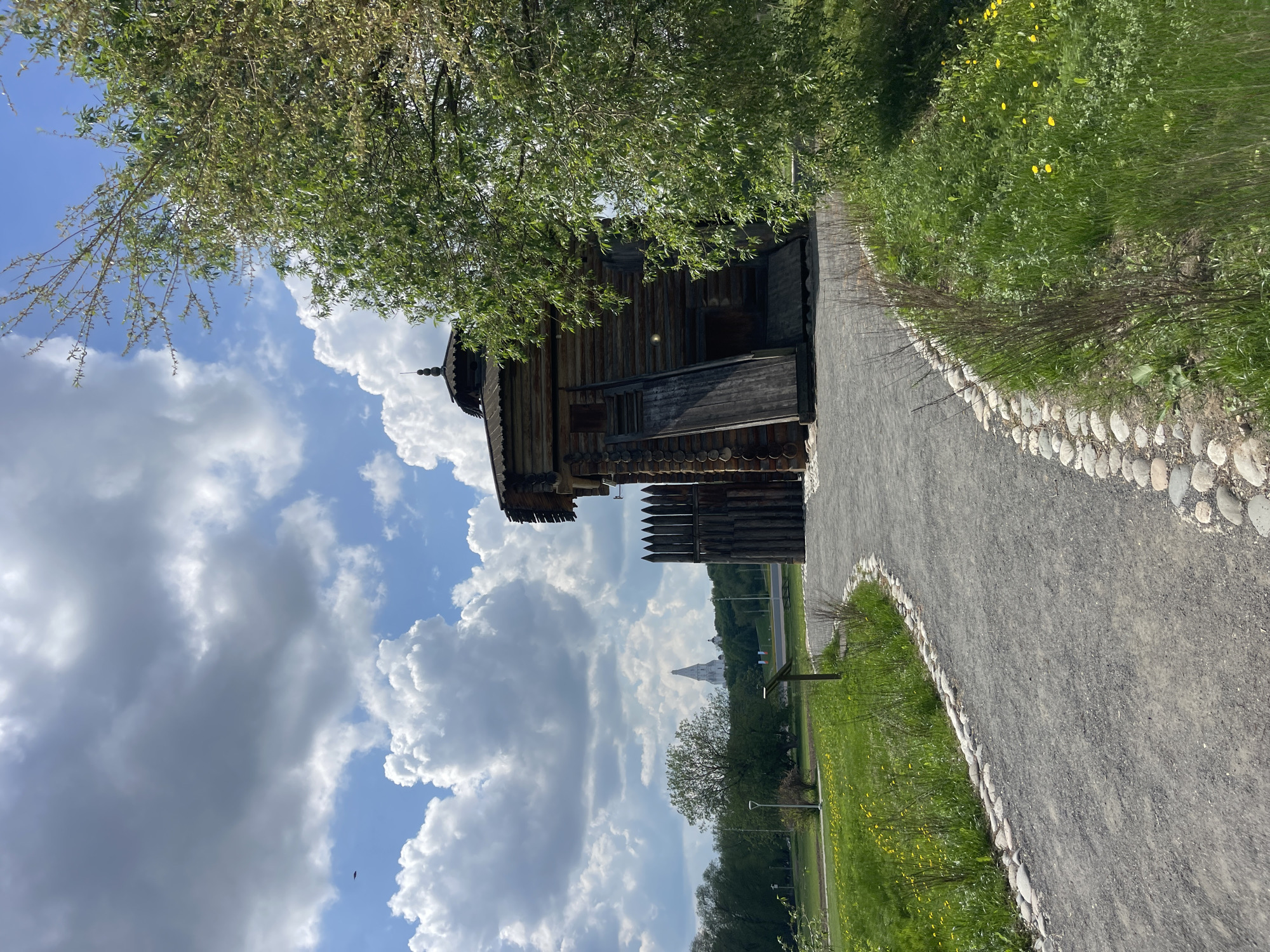 The image captures a scene in Kolomenskoye Park, a historical park located in Moscow, Russia. A wooden structure, likely a reconstructed or preserved historical building, stands adjacent to a pedestrian pathway. The pathway is made of gravel and edged with cobblestones. Lush greenery, including grass and yellow flowers, is visible alongside the path. In the background, beyond the wooden building, a light-colored church spire rises in the distance, obscured somewhat by trees. The sky is blue with large, white cumulus clouds. A tree is partially obstructing the view of the house and the sky.