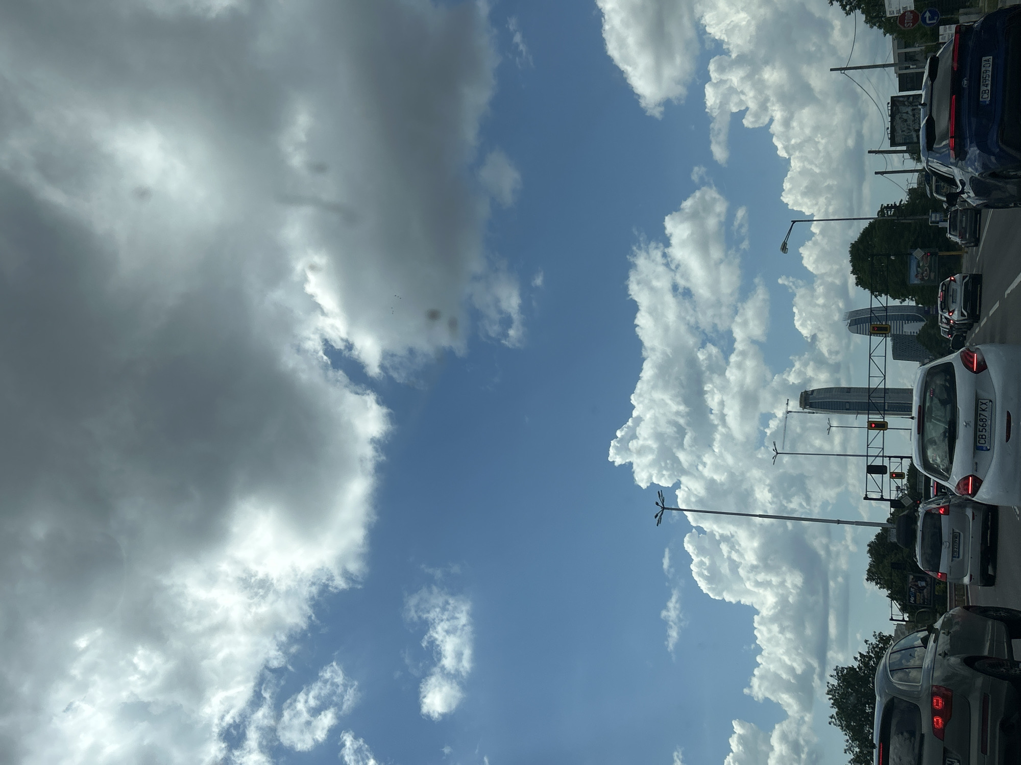 This image captures a daytime urban street scene in Sofia, Bulgaria, looking forward from a vehicle on a multi-lane arterial road, likely Tsarigradsko Shose. The sky dominates the upper half, featuring clear blue patches interspersed with large, bright white cumulus clouds and denser, darker grey clouds towards the top.
The foreground depicts multiple vehicles in moderate traffic, suggesting a stop or slow movement, likely due to the red traffic signals visible on the overhead gantry. On the far left, the rear-left quarter of a grey SUV is visible. In the center-right, a white Peugeot 208 with Bulgarian license plate "CB 5687 KX" is seen from the rear. To its right, partially visible, is the rear of a blue vehicle bearing license plate "CB 4959 OA". Further ahead in the midground, a grey Mazda with license plate "CB 2399 MB" is positioned.
Roadside infrastructure includes multiple tall streetlights with multi-arm fixtures, power lines, and a large metal gantry supporting traffic lights, which are illuminated red. On the far right, a red stop sign and a blue "turn right" arrow sign are visible.
In the background, beyond the trees lining the road, two distinct modern skyscrapers are visible: Capital Fort, a tall, slender building with a reflective facade, on the right, and Sky Fort, a similarly tall, curved, and tapering building, further to the left. A construction crane is faintly visible near Capital Fort. Green foliage from trees flanks both sides of the road, partially obscuring the distant skyline and buildings. No individuals are clearly discernible outside of the vehicles.