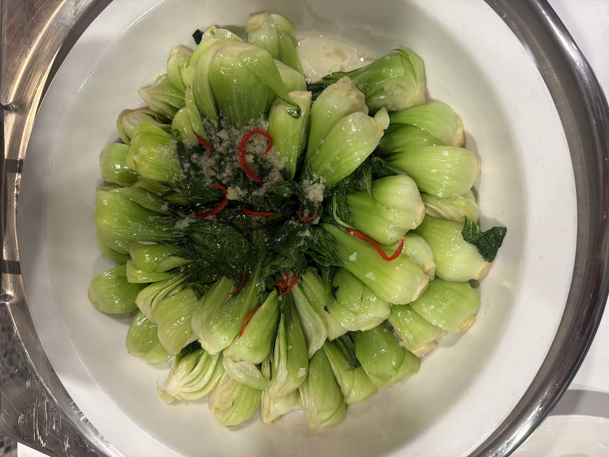 A top-down, eye-level photograph captures a large, oval-shaped white serving dish filled with cooked green vegetables, likely baby bok choy. The vegetables are arranged neatly in the dish, with numerous individual stalks visible. They feature light green stems and darker green leaves, appearing fresh and glossy, possibly coated in a light sauce or oil.
The center of the vegetable arrangement is generously garnished with finely minced white garlic and several thin, crescent-shaped slices of bright red chili peppers, providing a color contrast. A shallow pool of light-colored liquid is visible at the bottom of the dish, beneath the vegetables.
The white serving dish is positioned within a larger, metallic container, indicated by the shiny, silver-toned rim visible along the left and right edges. This metallic element suggests the food is presented in a buffet chafing dish or a similar food service setup. The lighting is bright and even, illuminating the food. There are no people, discernible text, or specific location cues beyond the buffet-style presentation.