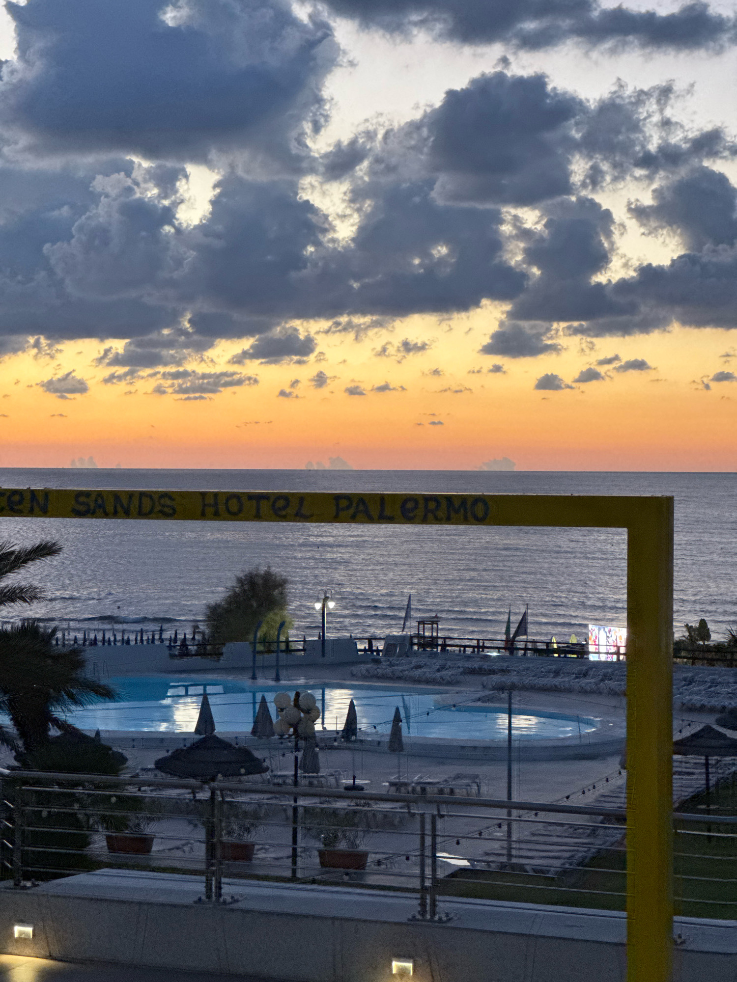 The image depicts a sunset scene at the "Golden Sands Hotel Palermo" in Isola delle Femmine, Italy. The sky is filled with dramatic, dark clouds against a vibrant orange and yellow sunset horizon. Below the sky, the calm sea stretches out, meeting the shore. In the foreground, a yellow structure with lettering visible spells out "GOLDEN SANDS HOTEL PALERMO". To the left of the yellow structure, a portion of a palm tree is visible. Further down, a swimming pool with a bright blue surface is illuminated, surrounded by lounge chairs and umbrellas. A railing with potted plants separates the viewer from the pool area. The scene appears to be from dusk, with artificial lights beginning to illuminate the resort area.