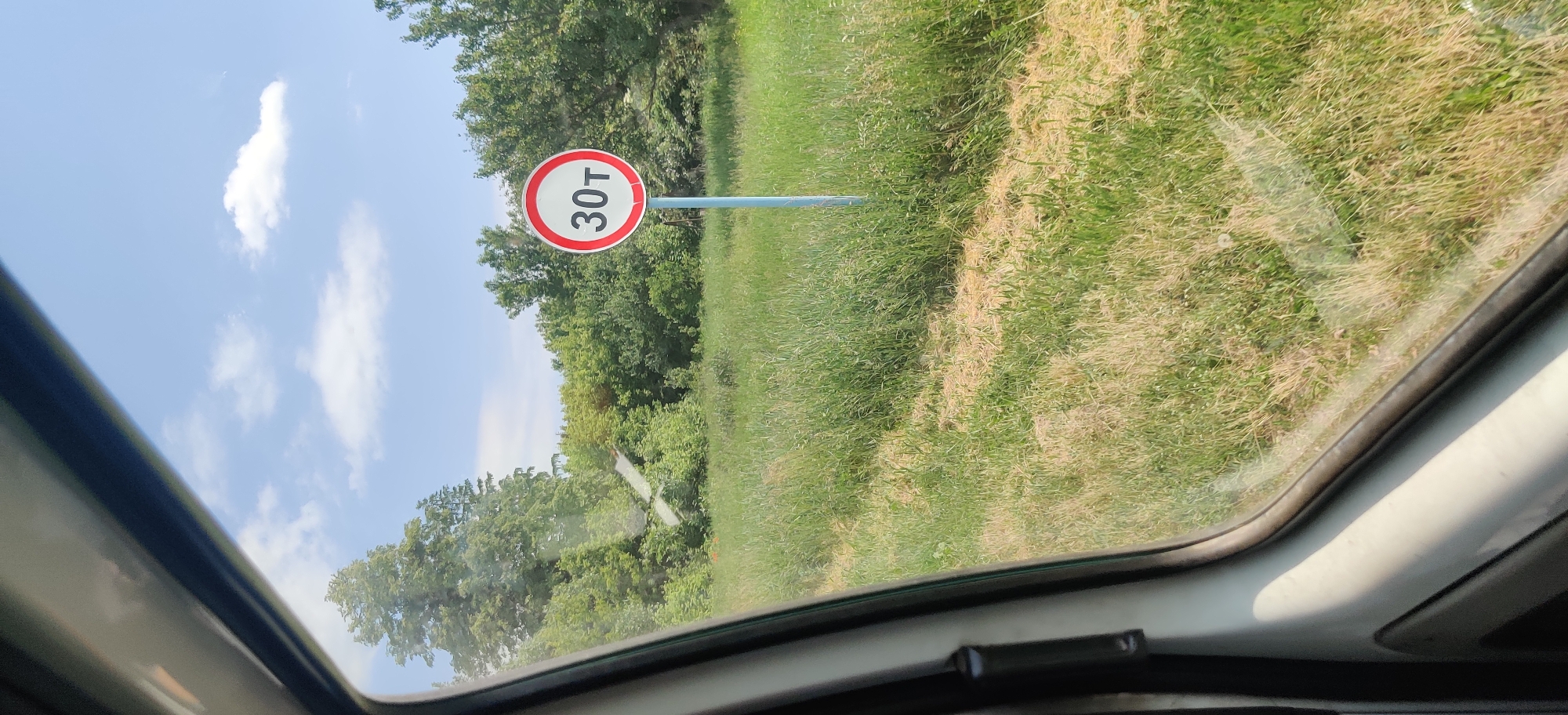 The image was taken from inside a vehicle, looking out the window. A traffic sign with "30т" in white text on a red circle is visible in the foreground, mounted on a blue pole. The sign indicates a weight limit of 30 tons. The background consists of a grassy field with scattered trees and a bright blue sky with some white clouds. The scene is set in Kolomak, Ukraine, as indicated by the location. The image does not contain any people or actions.