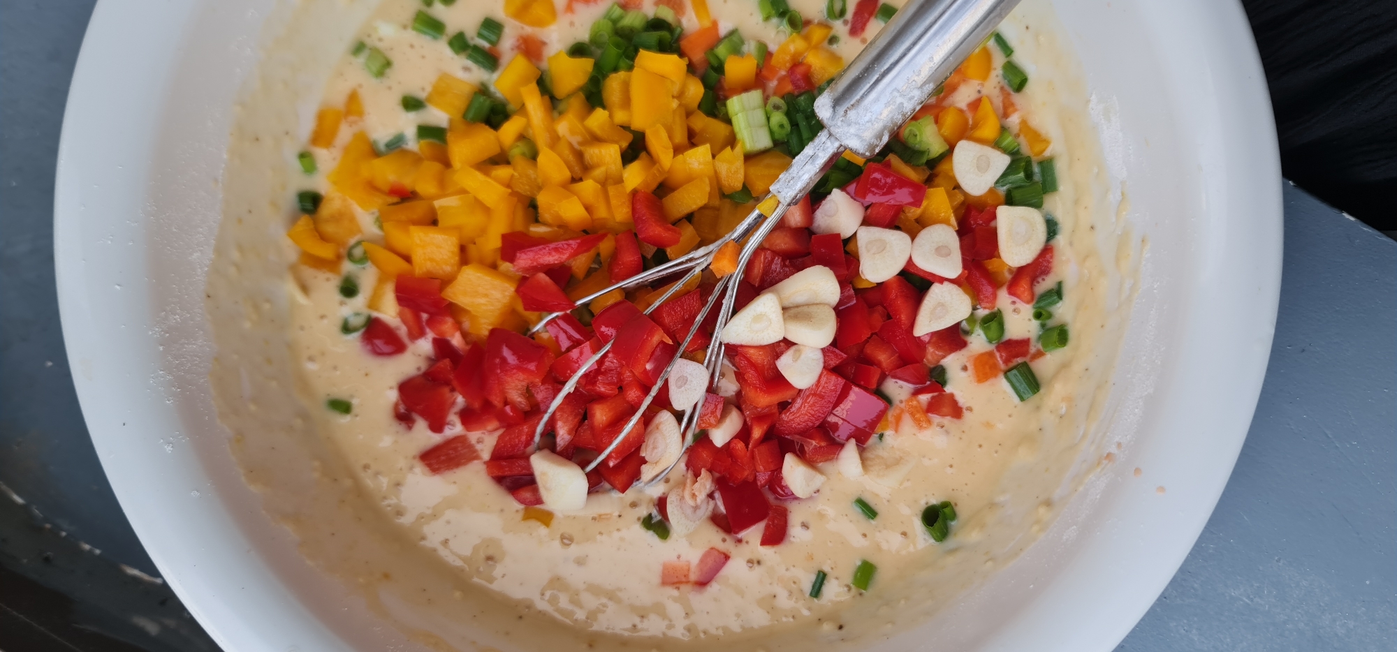 A top-down shot reveals a white bowl filled with a batter-like mixture and various chopped ingredients. Among the diced items are red and yellow bell peppers, green onions, and sliced garlic cloves. A wire whisk rests in the bowl, partially submerged in the mixture and intermingled with the ingredients. The bowl appears to be resting on a gray surface. The specific location within Đà Nẵng, Vietnam, is not discernible from the image.