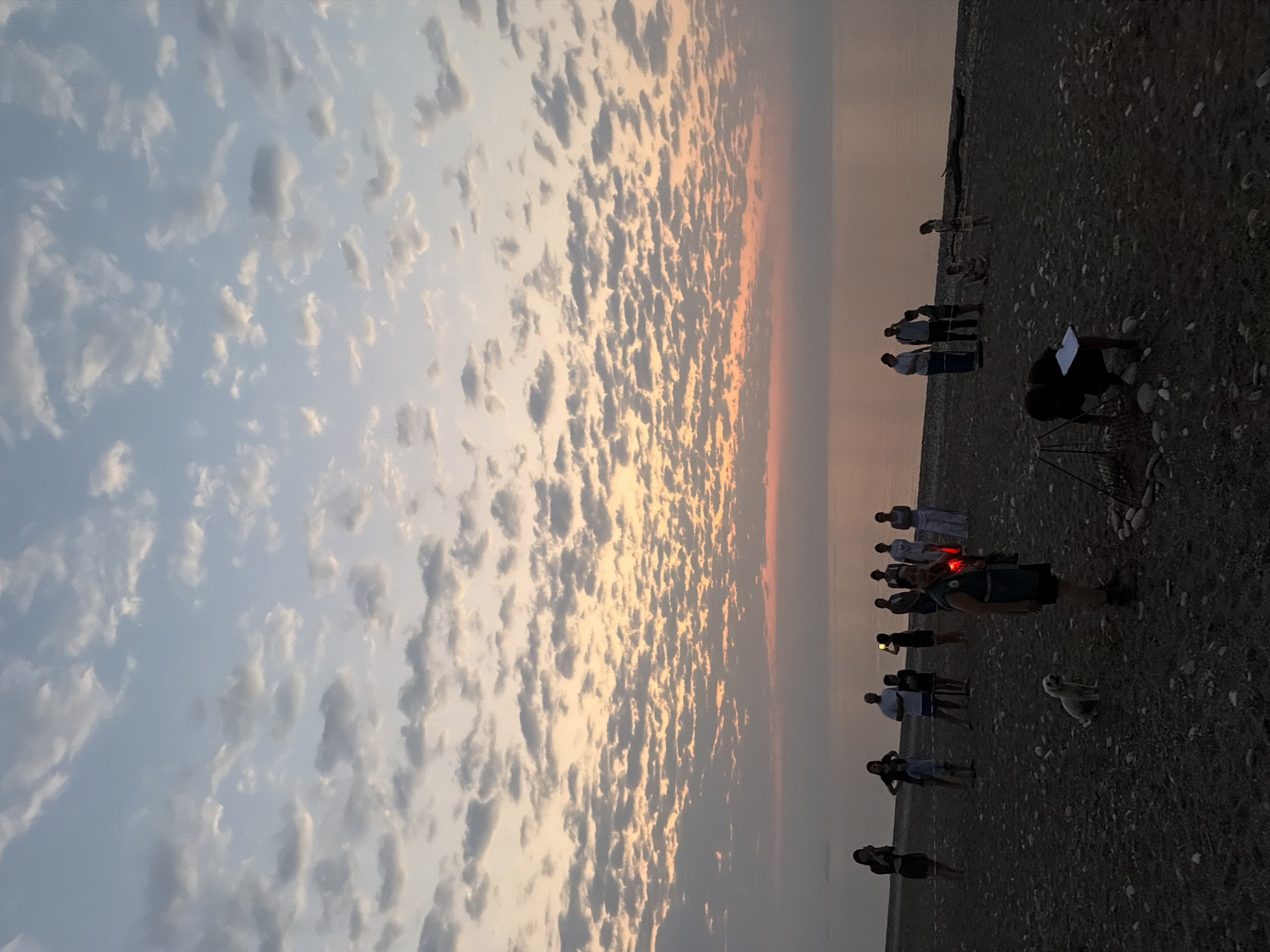 The image depicts a group of people gathered on a pebble beach, likely at sunset, with the sea and a cloudy sky as the backdrop. The location is specified as Kemer, Turkey.
In the foreground, there is a small white dog sitting on the pebbles. Further back, a person is crouched near a fire pit, possibly tending to a fire or preparing for a barbecue. This individual is holding a piece of paper or a notebook.
The main group of people is positioned between the foreground and the sea. They are standing and interacting, with some individuals holding up phones, perhaps taking pictures or videos. The group appears to be diverse in age, with adults and possibly children present. One person in the center of the group is wearing a dark vest and appears to be holding a lit object, possibly a flashlight or a lantern, which emits a red glow.
The environment consists of a dark, pebbly beach that slopes gently towards the calm sea. The sky above is filled with scattered clouds, illuminated by the soft colors of sunset, ranging from pale blue at the top to hues of orange and pink near the horizon. The water is smooth, reflecting the colors of the sky. There is a piece of driftwood on the beach in the middle ground.
The overall impression is one of a relaxed gathering on the beach during twilight. The presence of the fire pit and the group's engagement suggest a social event or a campfire activity.