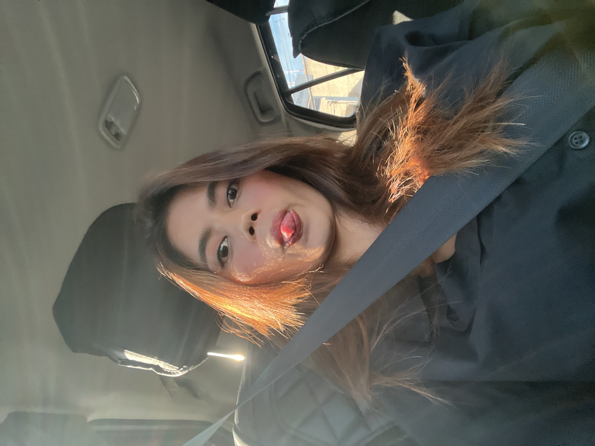 A woman is depicted seated in the front passenger seat of a vehicle, looking directly at the camera with her tongue extended in a playful gesture. Her long, auburn hair is brightly illuminated by strong sunlight entering from the left, creating a warm glow. She is wearing a dark-colored top and secured by a dark seatbelt. The car's interior features a light-colored headliner and an overhead light fixture. Through the rear passenger window, urban structures including a building with partially visible lettering "ERTIAN," utility poles, and overhead wires are discernible, consistent with an urban environment in Davao City, Philippines.