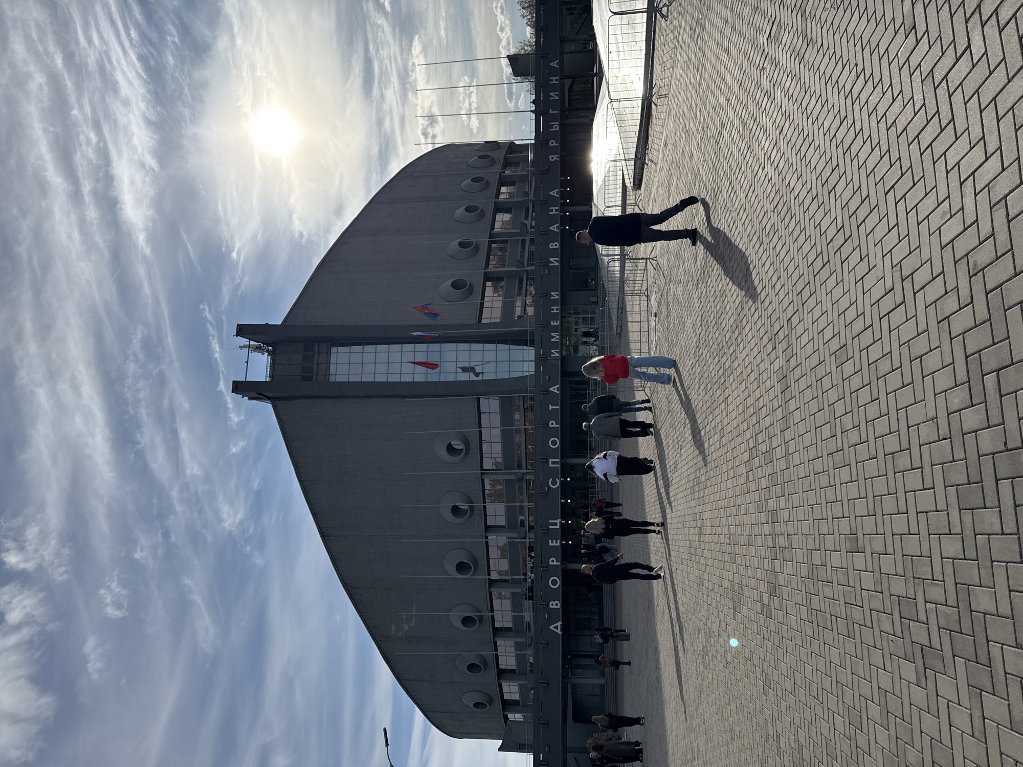 A photograph shows the Ivan Yarygin Sports Palace in Krasnoyarsk, Russia. The building is a large, curved structure with a gray exterior. There are several circular windows on the side of the building. The name of the palace is written vertically on the building in Cyrillic. Several people are standing in front of the building. The ground is paved with gray brick. The sky is blue with some clouds, and the sun is visible.