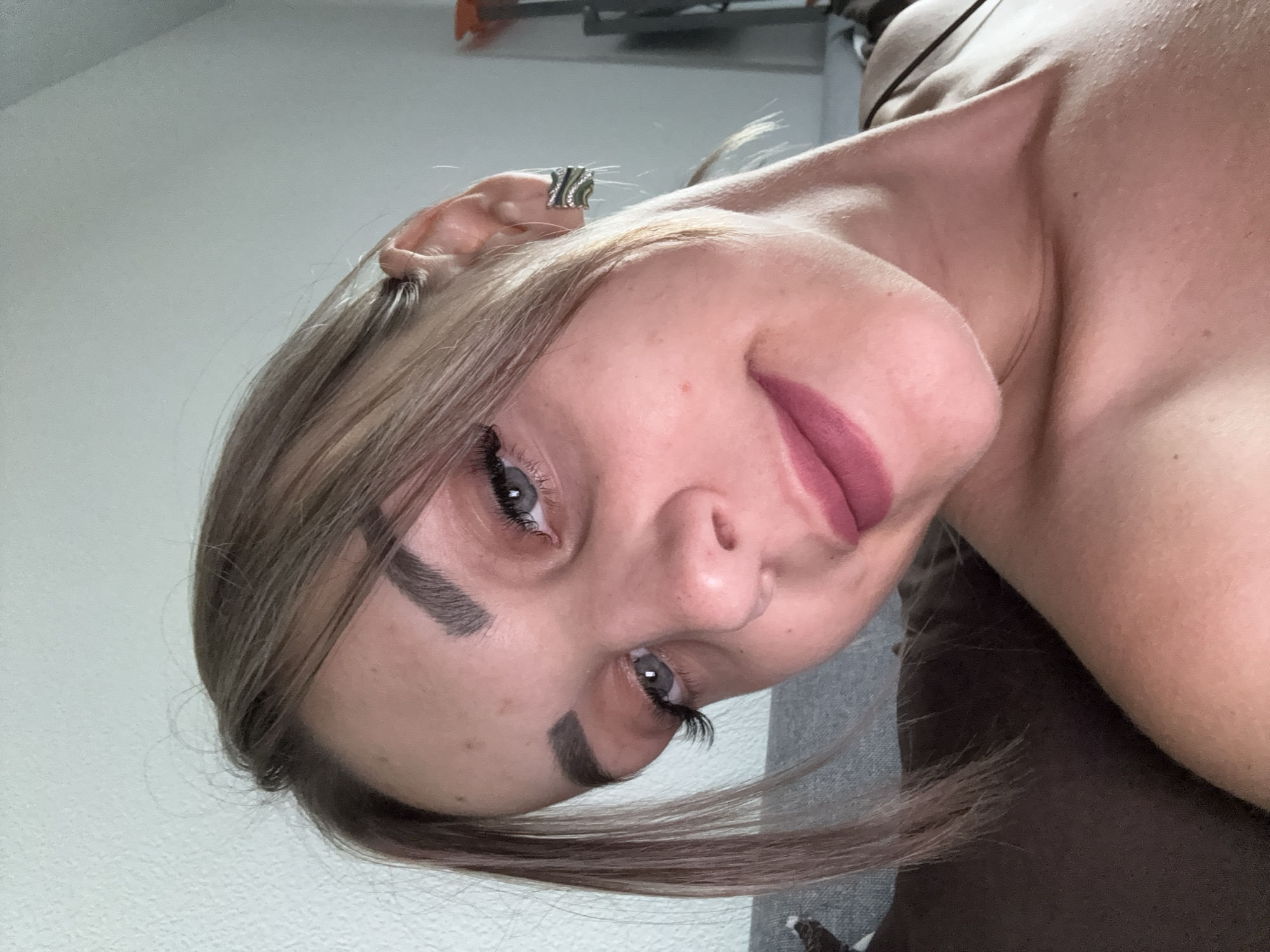 The image is a close-up, low-angle selfie of a young woman with blonde hair, blue eyes, and dark eyebrows. She is wearing makeup, including mascara, eyeliner, and lipstick. She has a silver and green earring in her right ear. Her skin is pale and has some freckles. She is leaning back on a brown surface that appears to be a couch or chair. The background is a white wall with some textured pattern. The lighting suggests an indoor setting, possibly a home. There are no discernible landmarks or context that would place the image in Yuzhno-Sakhalinsk, Russia.