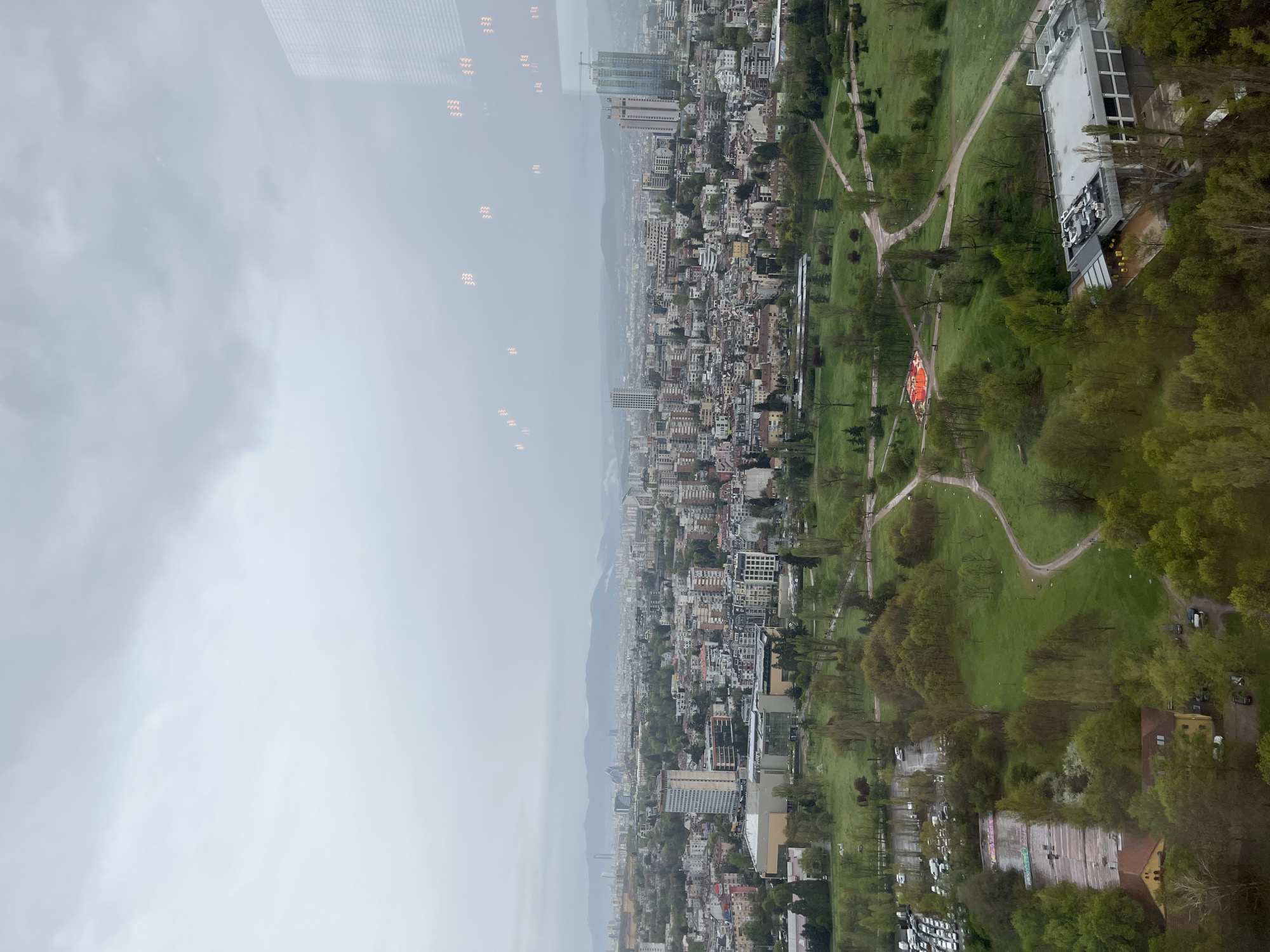 This high-angle image provides an expansive view of a city, identified as Sofia, Bulgaria, characterized by a large green park in the foreground blending into a dense urban landscape, with mountains visible on the distant horizon.
The park is lush with green grass and numerous trees, and is traversed by winding paved paths. Within the park, a prominent, brightly colored red-orange structure, possibly an artistic installation or play area, stands out. Adjacent to the park, a modern building with a light-colored roof and rooftop utilities is visible, along with a smaller, older building featuring a reddish roof and yellow walls towards the bottom right.
Beyond the green space, the city unfolds with a mix of buildings of varying heights and architectural styles, including multi-story residential blocks and taller structures, some appearing to be under construction with cranes visible. The distant mountains are partially obscured by atmospheric haze.
The sky is overcast and cloudy, casting a soft, diffused light over the entire scene, indicating daytime, likely a grey and humid day. Reflections of internal lights, appearing as wavy, orange patterns, are visible across the upper portion of the image, slightly overlapping the sky and distant cityscape. No discernible people or specific activities are clearly visible due to the distance. Very faint and unreadable white markings or graffiti can be seen on a surface at the bottom right edge of the park.