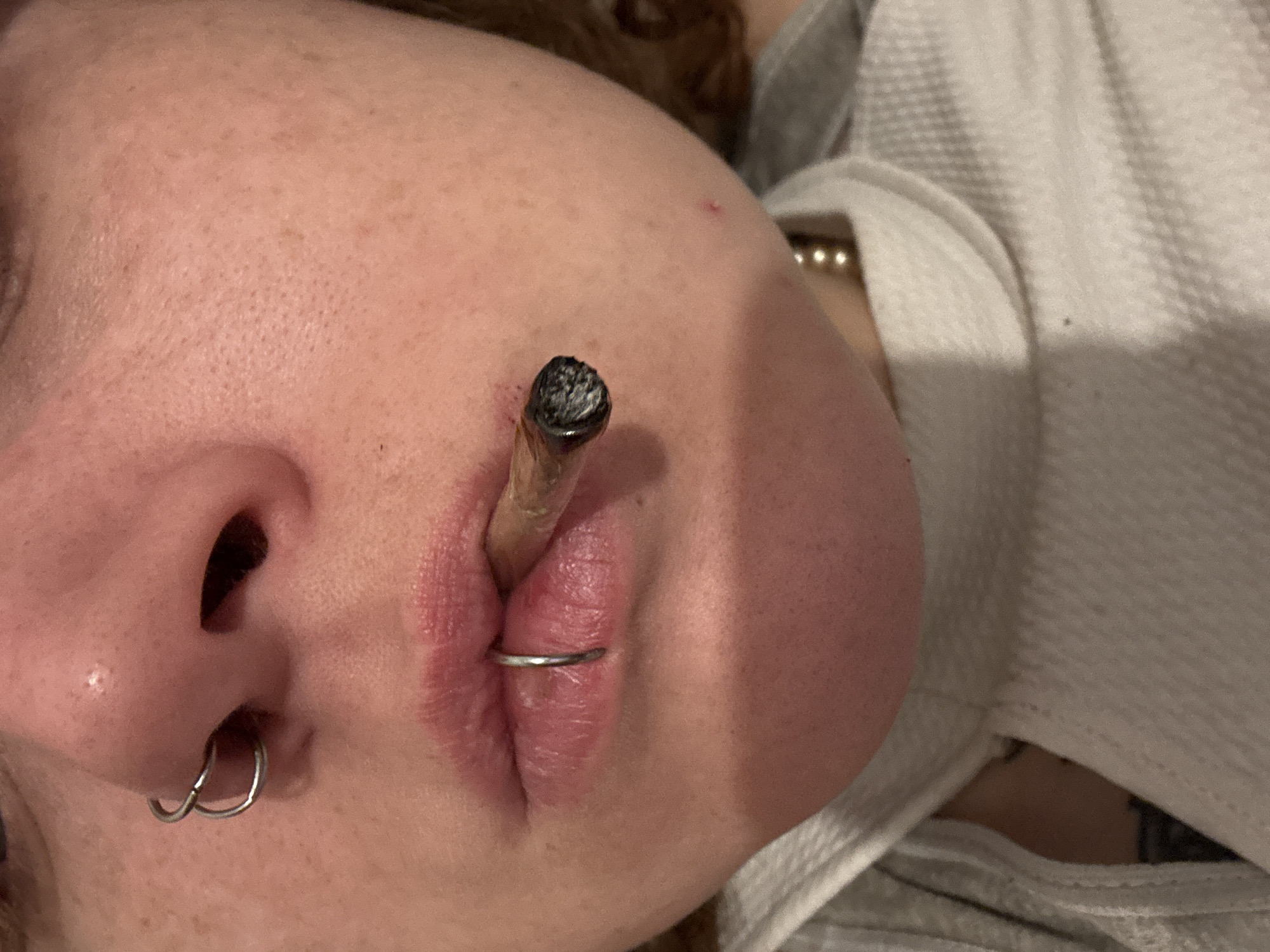 A close-up, vertically oriented photograph captures the lower half of a person's face and upper body in a semi-reclined position, likely indoors in Buenos Aires, Argentina. The main subject is an individual whose nose, mouth, chin, and a portion of their cheek and shoulder are visible.
The person is holding a partially smoked joint between their lips, with the tip heavily covered in ash, suggesting it is actively lit or recently used. Their lips appear a natural pinkish-red hue, showing some texture and possibly dryness or chapping. A silver-colored circular piercing is visible through their philtrum, below the septum. Another set of two silver-colored rings adorns their septum, piercing the base of their nose. The skin on their face and shoulder has visible pores, freckles, and a few small red blemishes.
To the right of the image, behind the person's head and shoulder, a light-colored, possibly white or off-white, textured fabric is seen, which appears to be clothing or bedding, perhaps a hooded garment or blanket. A faint glimpse of a gold-colored beaded necklace is visible on the person's neck/shoulder area, partially obscured by the fabric. The overall lighting is soft, creating an intimate and relaxed atmosphere. No text is visible in the image.