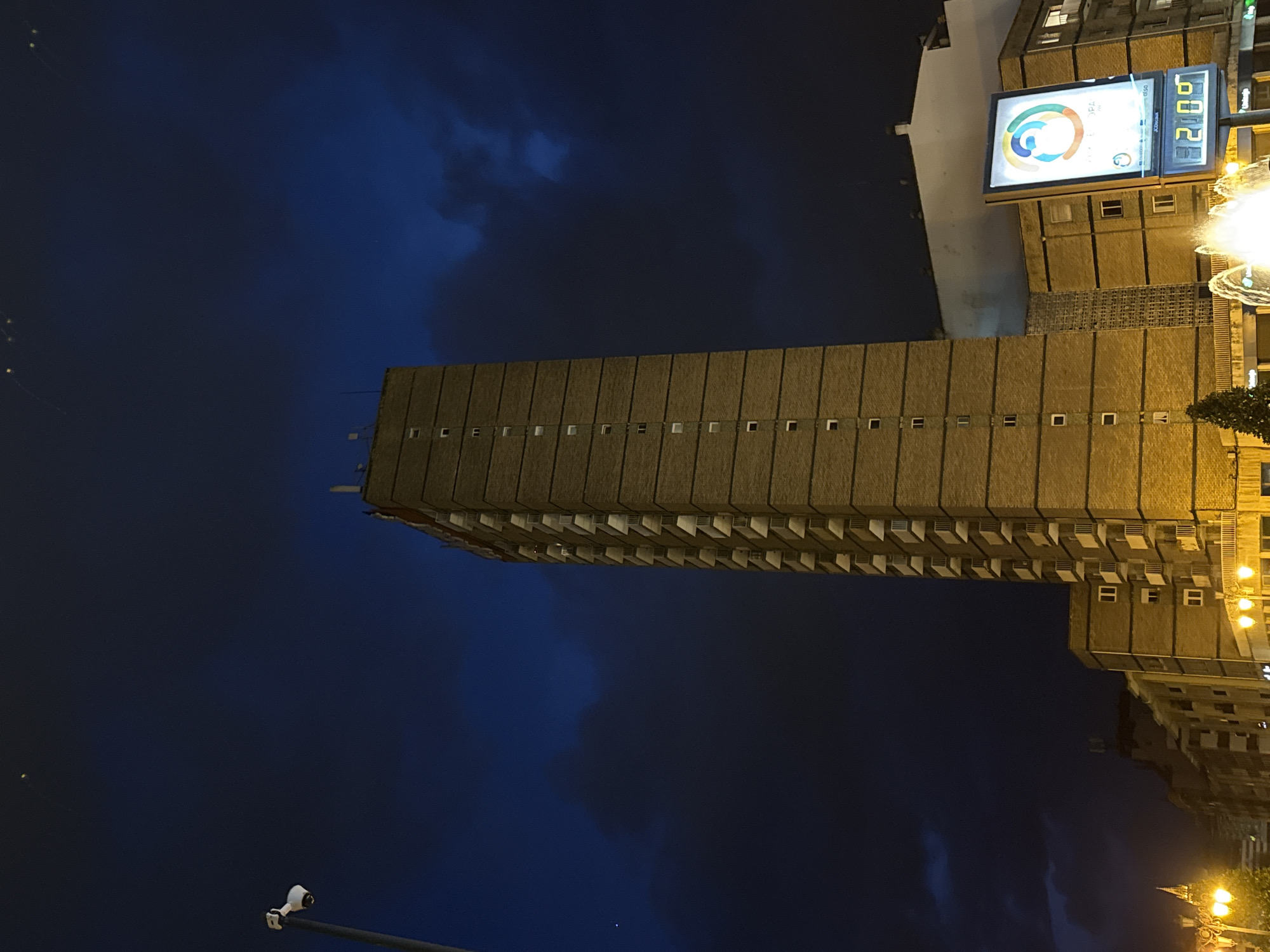 Here is a description of the image.
The image captures a tall, rectangular building under a dark blue evening sky, likely taken at night. The building is light brown or beige, with a repetitive pattern of small windows and horizontal lines suggesting multiple stories. To the right of the building, a large billboard displays what appears to be a colorful logo and some digital text, including the number "21:06". Streetlights and other buildings are visible in the background, indicating an urban setting. A security camera can also be seen in the bottom left corner.
Based on the presence of the San Lázaro building and the billboard, the location can be identified as Oviedo, Spain. Specifically, the tall building is the Residencial San Lázaro. The billboard is near the intersection of Avenida de Colón and Calle Samuel Sánchez, indicating the image was taken from that vicinity.
