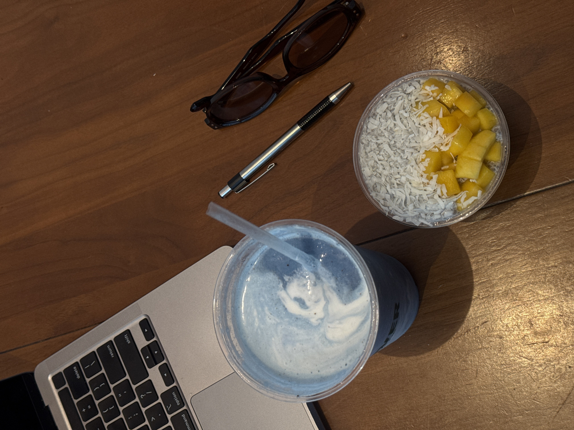An overhead shot captures a collection of items arranged on a light brown wooden surface, likely a table or desk, in an indoor setting. The scene suggests a work or study break.
In the lower left, a silver laptop keyboard is partially visible, showing keys such as "delete," "return," "shift," "option," and "command." Immediately to its right is a clear plastic cup containing a light blue beverage with white foamy swirls on top, and a clear straw inserted. The cup is encased in a blue holder or sleeve, on which some indistinct text ending with "R" is partially visible.
Above the laptop and blue drink, a pair of dark sunglasses with brown tinted lenses rests, folded. Small white text reading "MADE IN CHINA" is discernible on one of the sunglass arms. Next to the sunglasses, a silver pen with a black textured grip and a pocket clip is laid out.
In the upper right quadrant of the image, a clear plastic container holds a snack or dessert. The contents are divided, with one half consisting of white shredded coconut and the other half filled with small, diced yellow mango pieces, likely part of a chia pudding or fruit bowl.
The lighting creates some soft shadows, indicating an indoor environment with ambient light. No people are visible. Given the location context of New York, United States of America, the scene depicts a typical urban work or leisure setting.