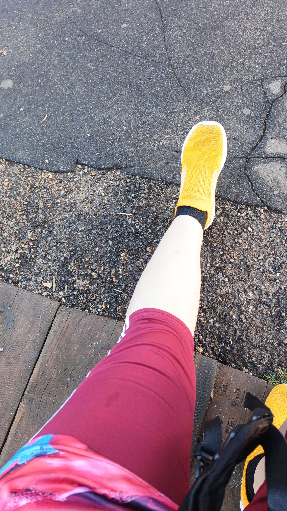 The image shows a person's legs and feet from a top-down perspective. The person is wearing burgundy athletic shorts with white stripes, a colorful patterned shirt partially visible, a yellow sock, and a yellow shoe. The ground appears to be a mix of asphalt and gravel, with some wooden planks in the foreground. The context provided of Drezna, Russia, is external information and not discernible from the image itself. The scene is outdoors, likely in a park or recreational area given the materials.