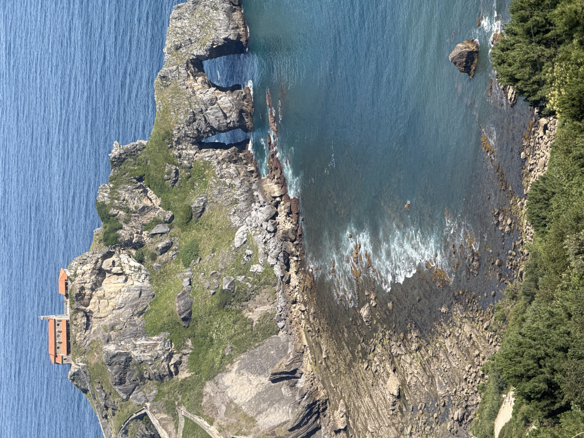 The image shows the San Juan de Gaztelugatxe hermitage in the municipality of Bermeo, Basque Country, Spain. The hermitage is situated on a rocky islet connected to the mainland by a stone bridge. The islet is steep and covered in grass and rocks. Two large natural arches are visible in the rocky coastline. The sea is a deep blue, with waves crashing against the shore. In the foreground, there are trees and a walking path. The image is taken from a high vantage point, offering a panoramic view of the landscape. There are no people visible in the image.
