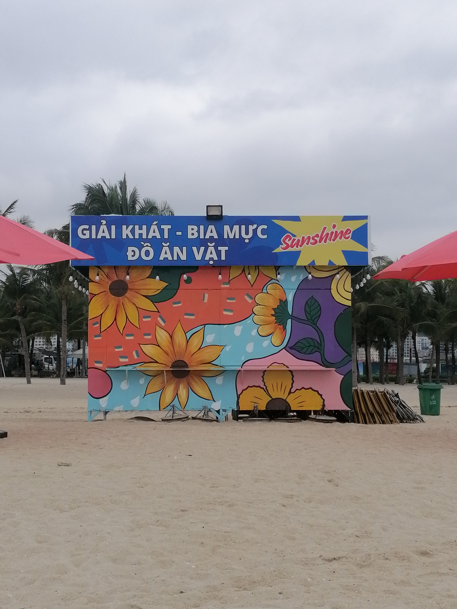The image displays a colorful vendor stand situated on a sandy beach, likely in Bai Chay Ward, Vietnam. The sky is overcast, indicating a cloudy daytime. No people are visible in the scene.
The central subject is a vibrant, rectangular stand, brightly painted with large, stylized sunflowers and abstract designs in shades of orange, light blue, purple, pink, and yellow. A string of globe lights hangs along its upper edge. Above the painted stand is a blue sign with white text that reads "GIẢI KHÁT - BIA MỰC ĐỒ ĂN VẶT," which translates to "REFRESHMENTS - BEER SQUID SNACKS." To the right of this text, within a yellow starburst shape, the word "Sunshine" is written in a cursive font. A single spotlight is mounted on the top of the blue sign.
To either side of the stand, portions of large red beach umbrellas are visible. The background features numerous palm trees, with some distant buildings visible through the foliage, suggesting a developed coastal area. On the right side of the stand, a green trash can and a pile of stacked wooden or metal poles, possibly folded beach chairs or umbrellas, are present. The foreground consists of light brown sand, showing faint indentations. The overall setting is a typical beachside concession, ready to serve visitors refreshments and snacks.