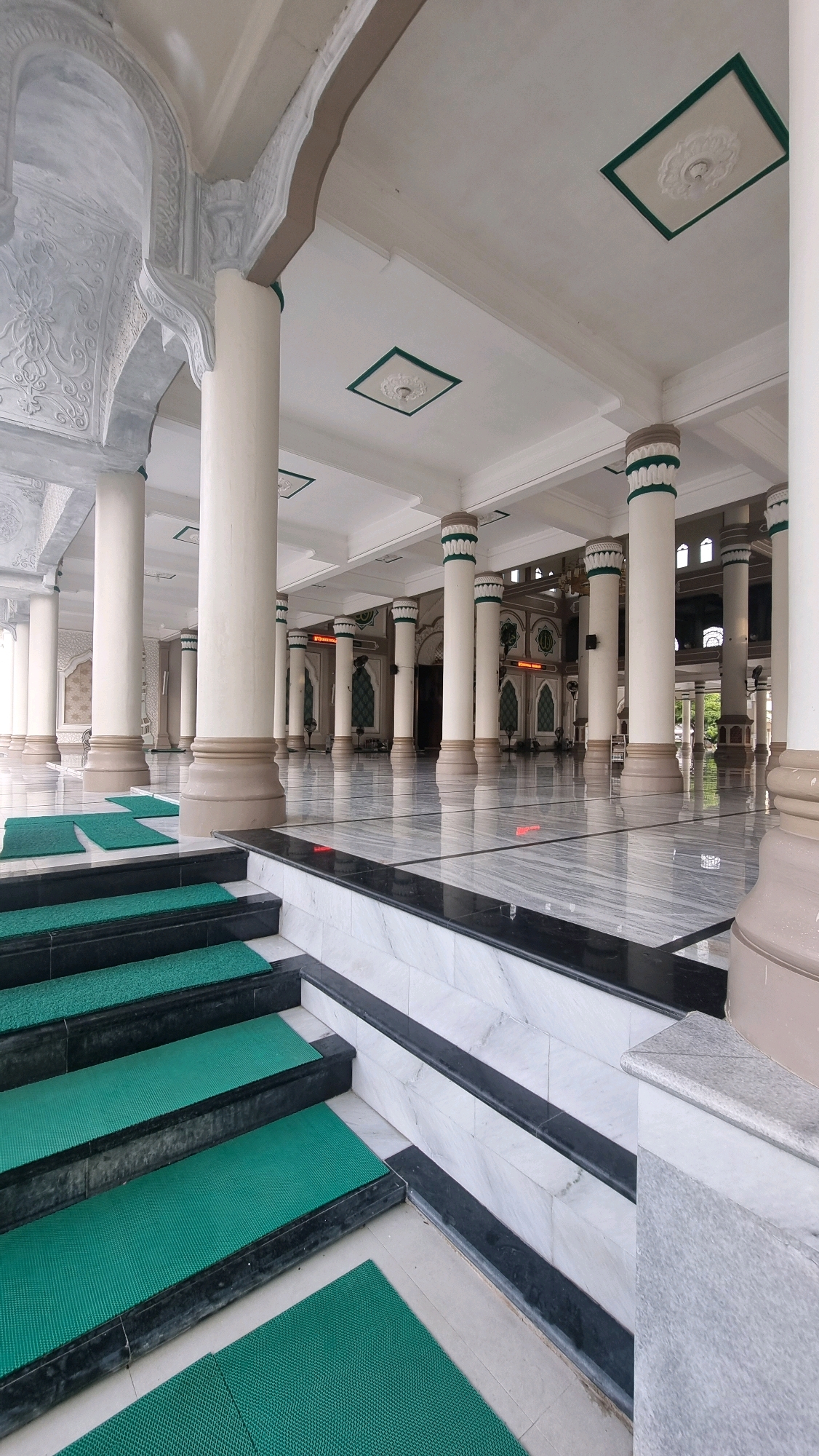 This is a full-shot, eye-level, indoor, vertical, and wide-angle photograph of the interior of the Islamic Center Lhokseumawe, located in Lhokseumawe, Indonesia. The spacious hall features a polished marble floor that reflects the light from the ceiling. Rows of tall, white, cylindrical columns with decorative green and white accents line the hall, supporting a white ceiling with square patterned recesses. A set of black marble stairs with green mats leads up to the main hall. Intricate white relief carvings adorn the arches and structural elements on the left side of the image. The overall impression is one of grandeur and serenity.