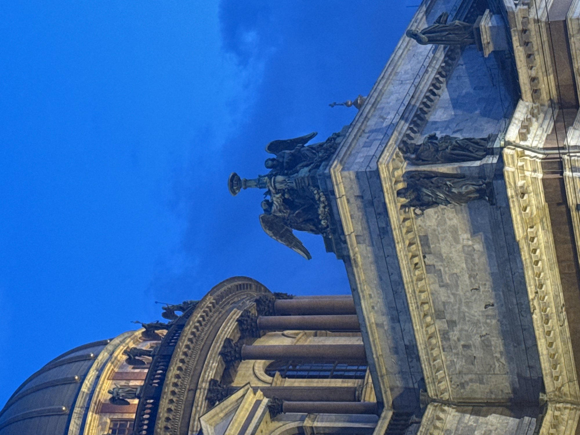 The image captures a dramatic, diagonally oriented, low-angle view of an ornate architectural structure in Saint Petersburg, Russia. Taken during twilight or night, the building is illuminated by warm artificial lights against a deep blue sky with subtle cloud formations. A large, dark-colored sculpture of an angel with prominent wings, holding an ornate censer or candelabra, is prominently featured on a building corner. To its left, a segment of a vast, ribbed dome is visible. To the right, the building's facade showcases multiple columns and further elaborate sculptural figures, including reclining and standing statues along the cornices. The stone facade glows, contrasting with the cool blue of the sky and the dark tones of the bronze sculptures. No discernible text is visible, and no specific activity is taking place. The scene conveys historical grandeur and intricate artistry.