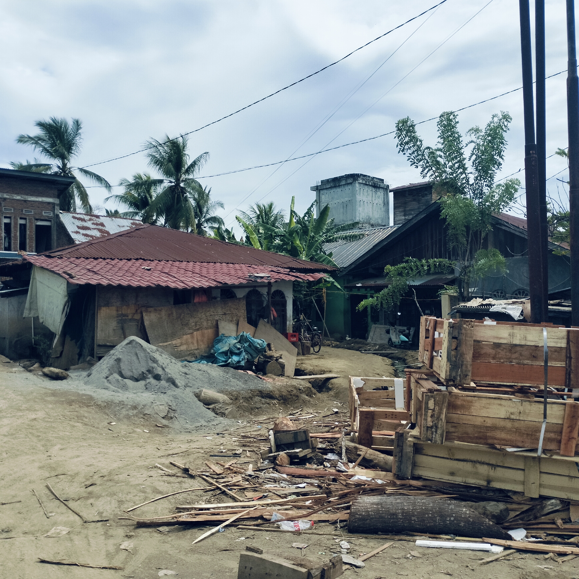 This outdoor scene, set in Beurawang, Indonesia, depicts a bustling residential area under an overcast sky. The foreground is dominated by unpaved, dusty ground littered with various pieces of wood, construction debris, and a large fallen log. On the left, a substantial pile of sand or gravel suggests ongoing construction or renovation, with blue tarps or bags nearby.
Several houses and structures are visible throughout the image. To the left, a single-story dwelling with a prominent red tiled roof appears somewhat makeshift, with plywood and other materials leaning against its white-washed walls. Behind it, a brick building with visible window frames is partially obscured. Further into the mid-ground, a dark, wooden-sided house with a dark roof stands, and further back and to the right, a tall, grey concrete structure, possibly a water tower or a swiftlet house, rises above the rooftops.
Lush tropical vegetation is abundant, with several tall palm trees (likely coconut palms) dotting the landscape, especially towards the left and center-background. Banana plants are also visible behind some of the houses. Electrical wires crisscross the sky, connecting to several tall, dark utility poles on the far right of the frame.
A bicycle is parked near the red-roofed house, and a small scooter or motorcycle is visible further back on a dirt path. Among the scattered debris in the foreground, wooden crates or pallets are stacked, adding to the impression of a working or developing area. A red Coca-Cola crate is visible near the bicycle. The scene captures a typical daytime view of a community in a tropical setting, characterized by mixed housing styles, natural elements, and everyday clutter. No specific human activity is clearly depicted, suggesting a quiet moment in a lived-in environment.