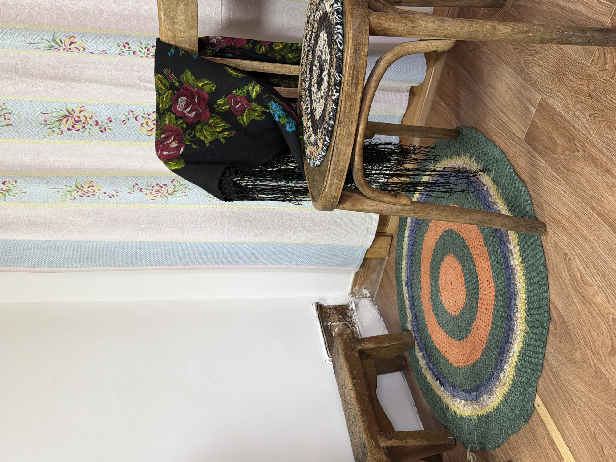 In a dimly lit room, likely in Ust'-Pyzas, Russia, a rustic wooden chair stands as the focal point. A black shawl with a vibrant floral pattern of red roses and green leaves is draped over its back. The shawl is edged with black fringe that hangs down. On the seat of the chair rests a small, circular braided rug, predominantly black and white with brown accents. Beside the chair, a simple wooden stool, worn and aged, is positioned near a wall. A circular rag rug with concentric rings of green, orange, purple, and yellow lies on the wooden floor. The wall behind the objects is painted white, and a set of pastel-colored curtains, featuring stripes and floral motifs, hangs to the right. The floor is made of wooden planks. The scene evokes a sense of traditional rural living.